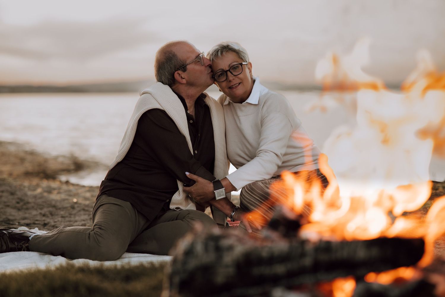 Elderly couple sharing a romantic kiss by a beach campfire at sunset, enjoying a cozy outdoor moment together.