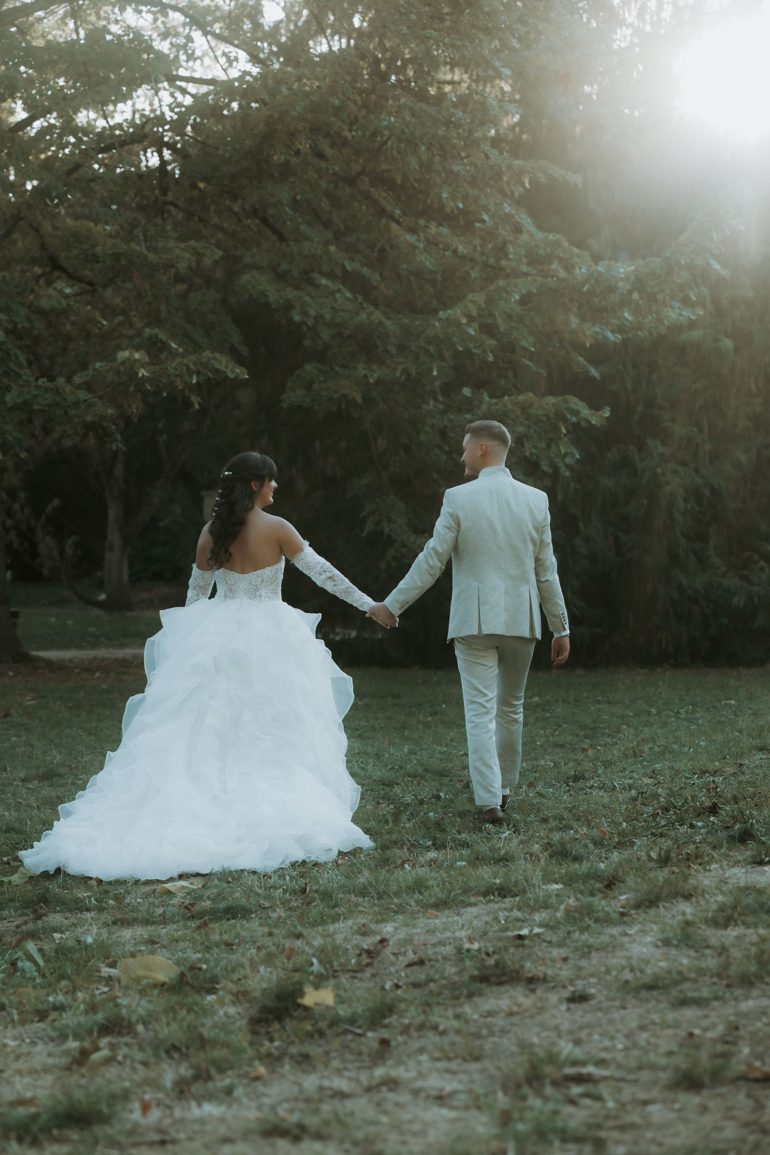 Bride in white gown and groom in beige suit walk hand in hand through lush green park in golden sunlight.