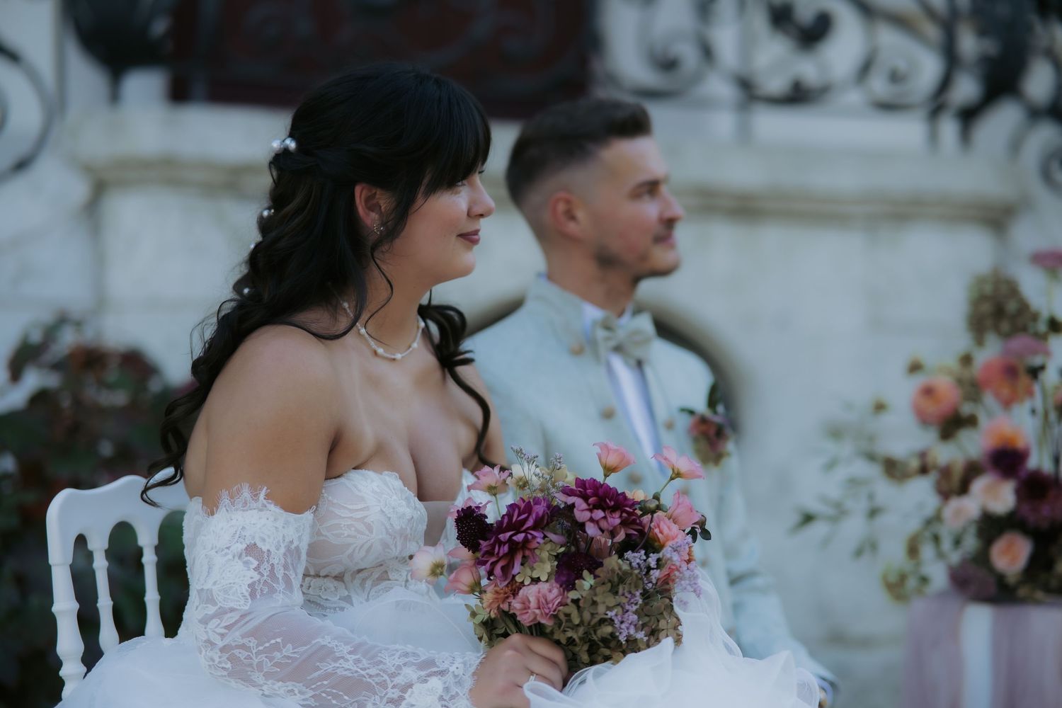 Bride in white lace gown holding floral bouquet sits beside groom in light suit at outdoor wedding ceremony.