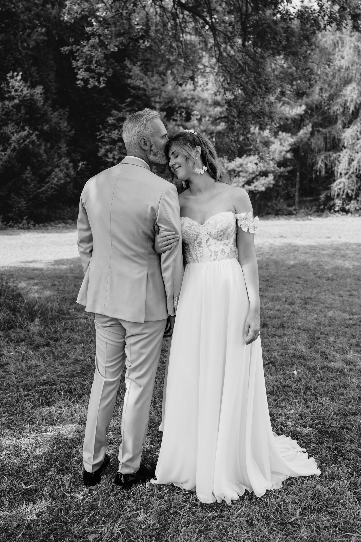 Black and white photo of a bride and groom sharing a tender moment outdoors, surrounded by lush trees.