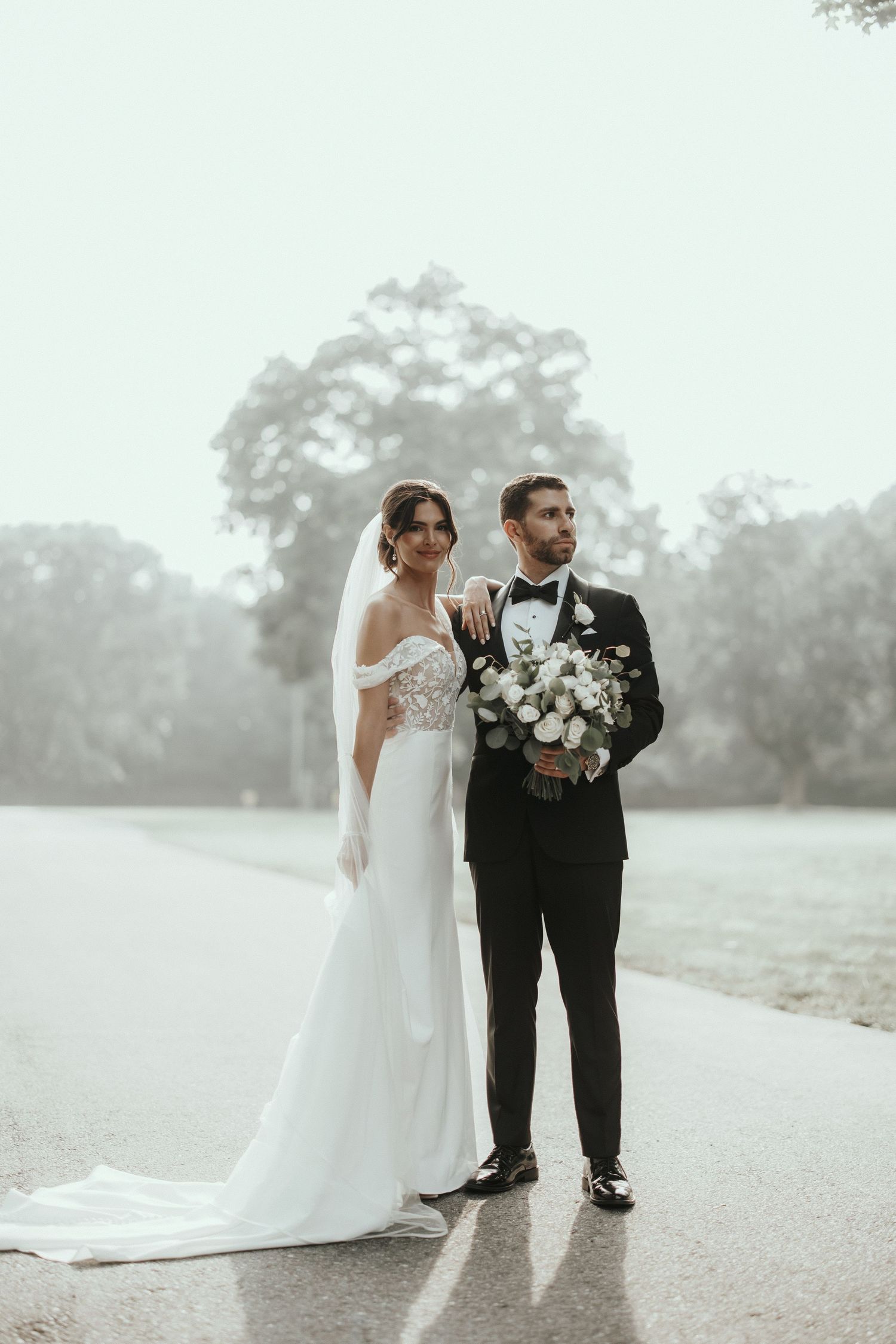 Elegant bride and groom share a moment on a misty pathway in formal wedding attire.