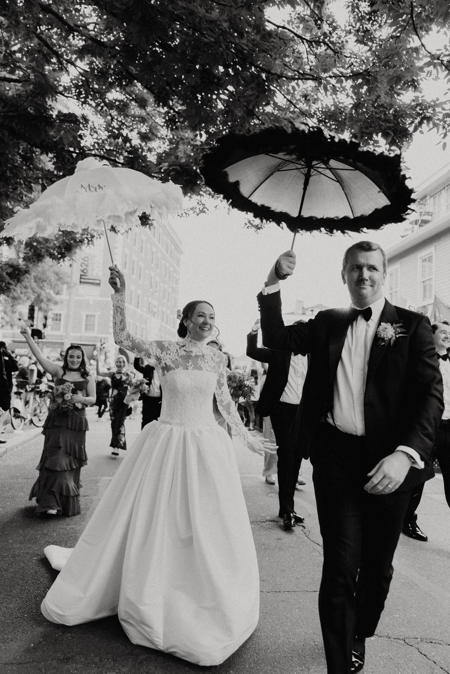 Black and white photo of a bride and groom holding decorative umbrellas during a wedding parade on a city street.