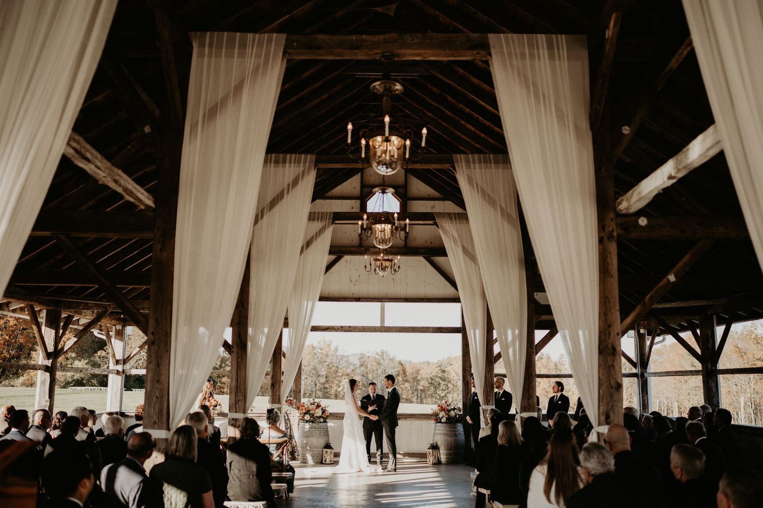 A rustic barn wedding ceremony with white draping, chandeliers and guests seated in rows beneath wooden beams.