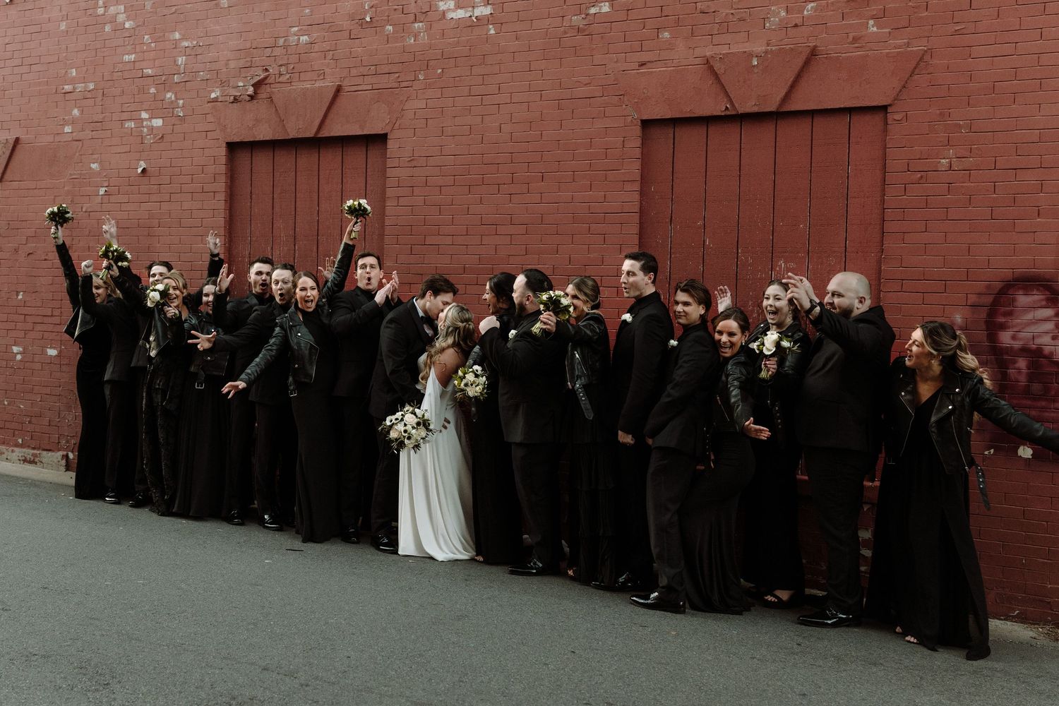 Wedding guests line up against red brick wall for group photo.