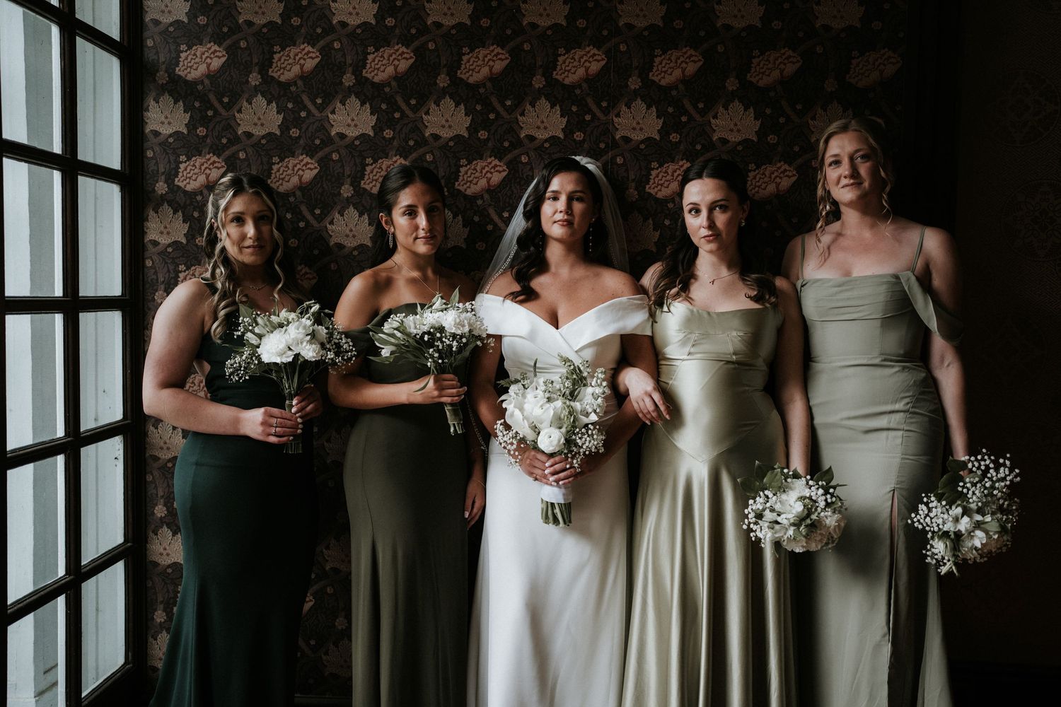 Wedding party poses together in elegant olive green dresses and white gowns by large windows.