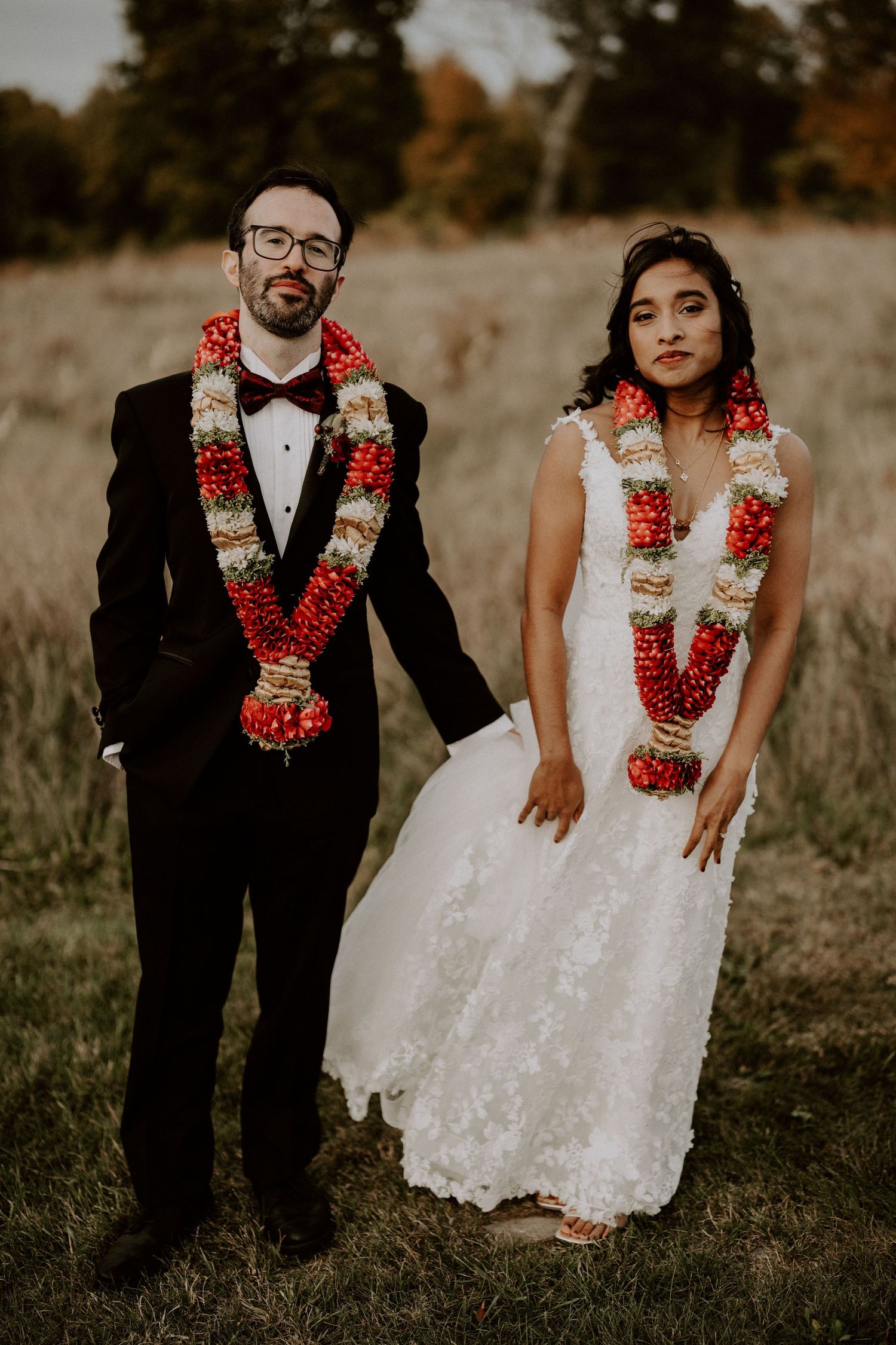A couple wearing traditional wedding attire with bright red and white floral leis pose together in a grassy field.