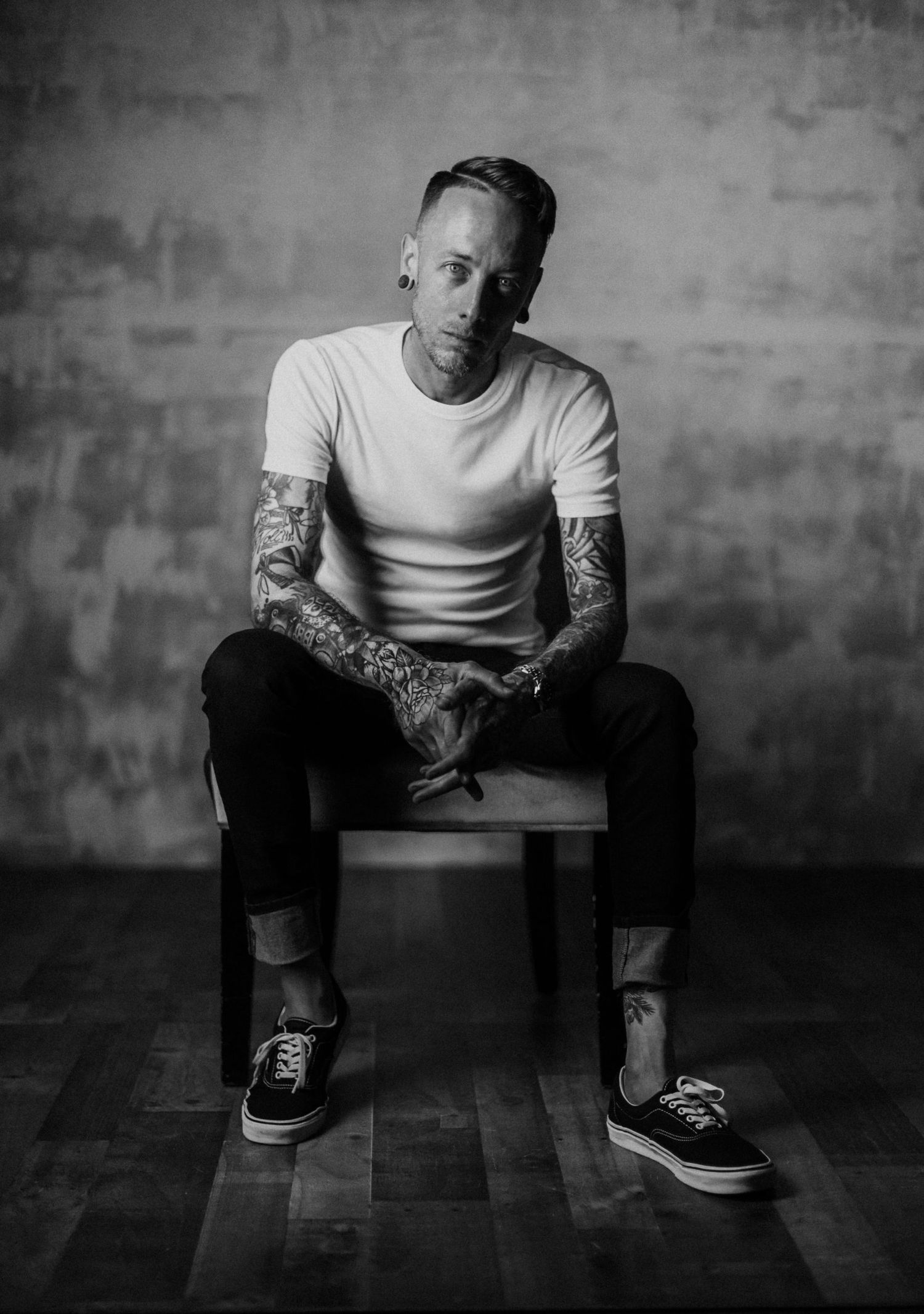 Artistic black and white portrait of a tattooed man in a white t-shirt sitting on a stool.