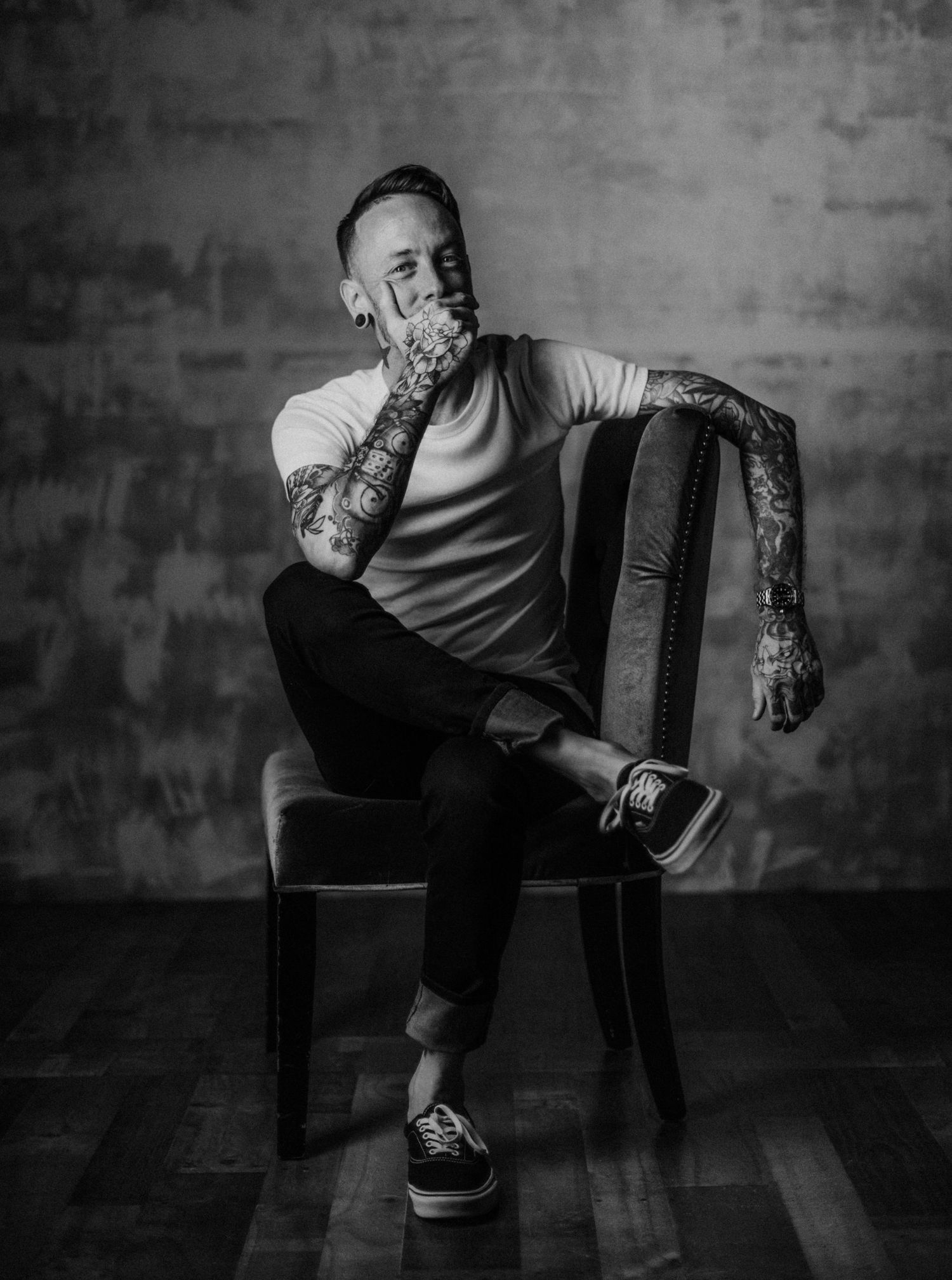 Moody black and white portrait of tattooed person seated on chair against concrete wall.
