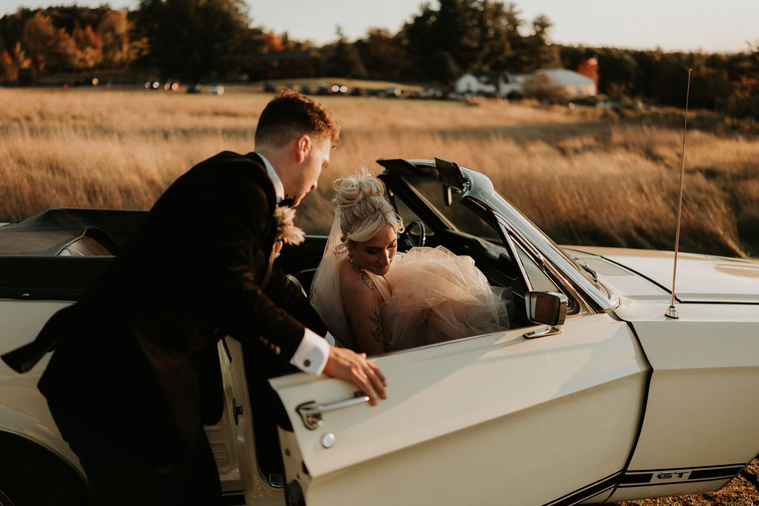 A vintage car parked in a golden field during sunset creates a romantic wedding day atmosphere.