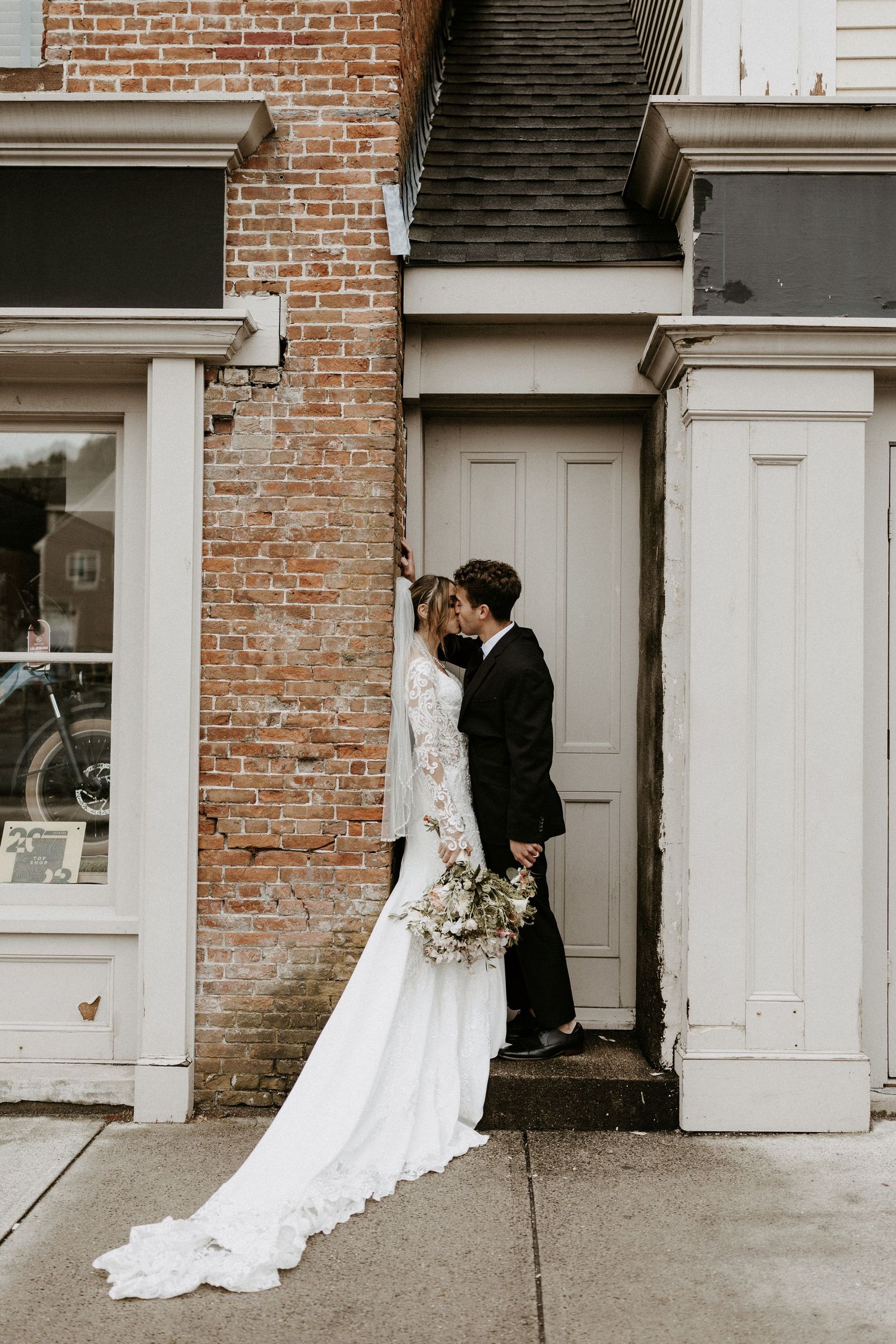 A couple shares a romantic moment against a brick wall with a flowing white wedding dress.
