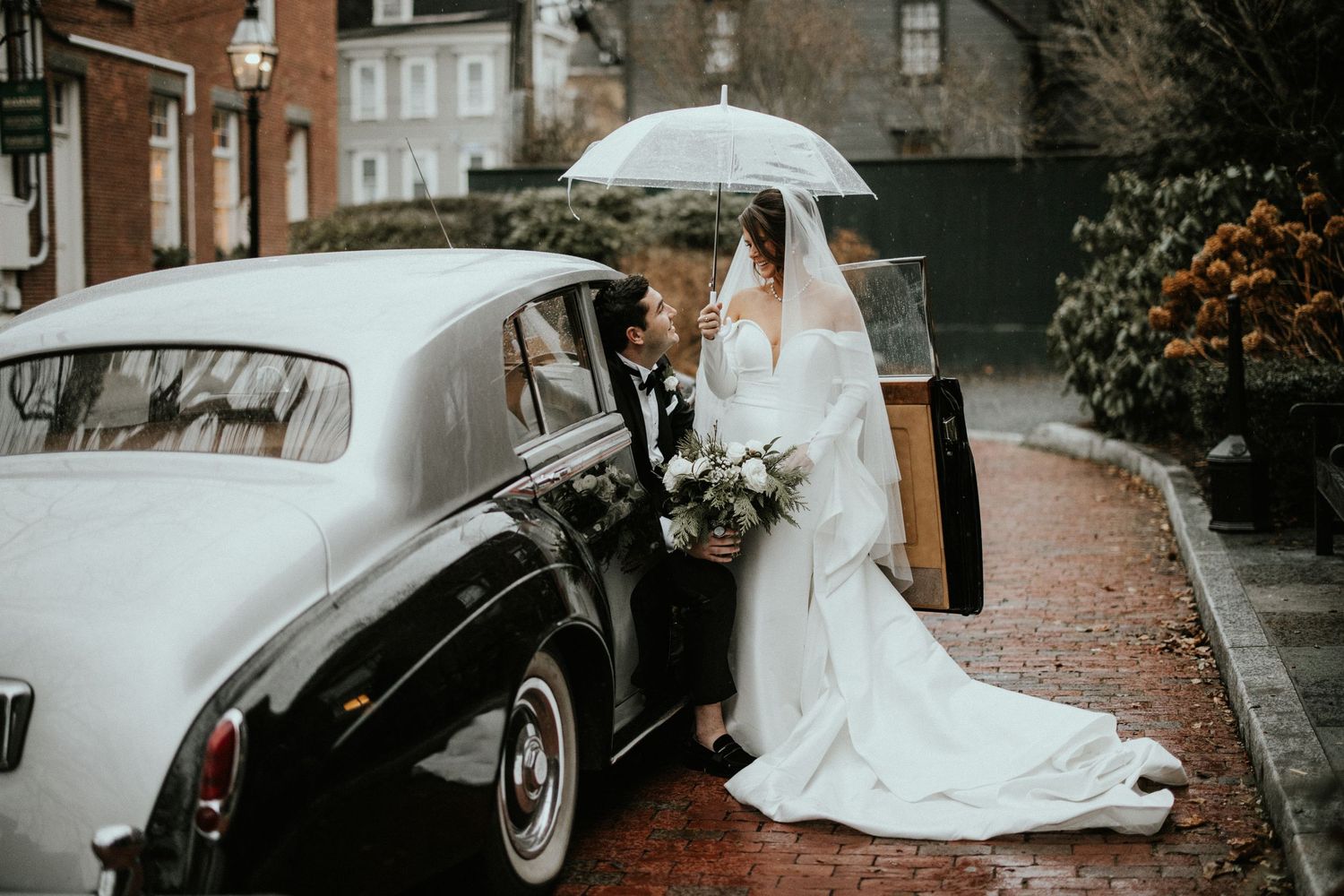 A rainy wedding day scene with a white umbrella and elegant bridal gown against brick buildings and vintage cars.