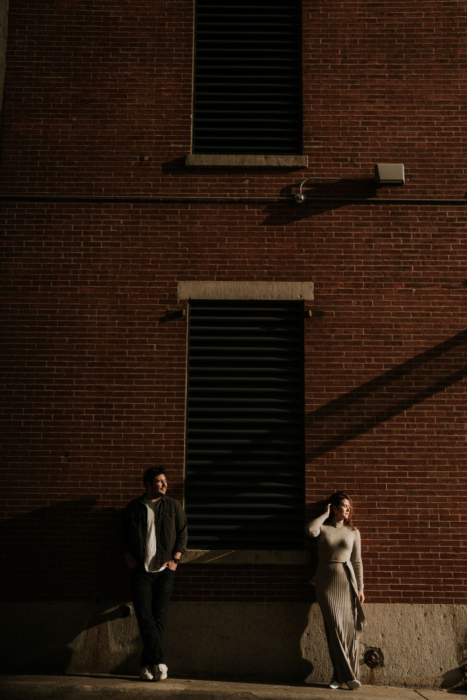 A couple sits against a brick wall in dramatic lighting.