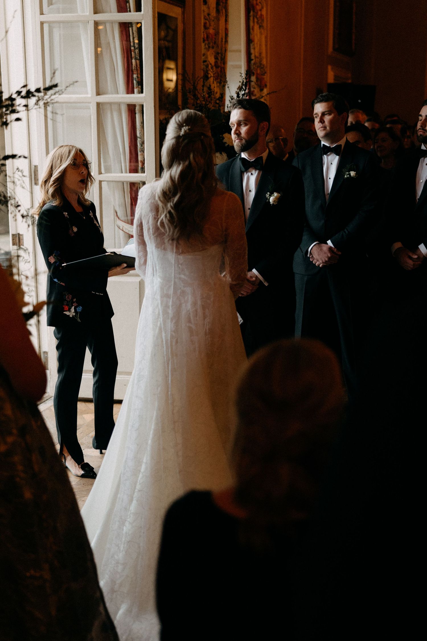 Bride and groom exchange vows during elegant indoor wedding ceremony with officiant and groomsman standing nearby.