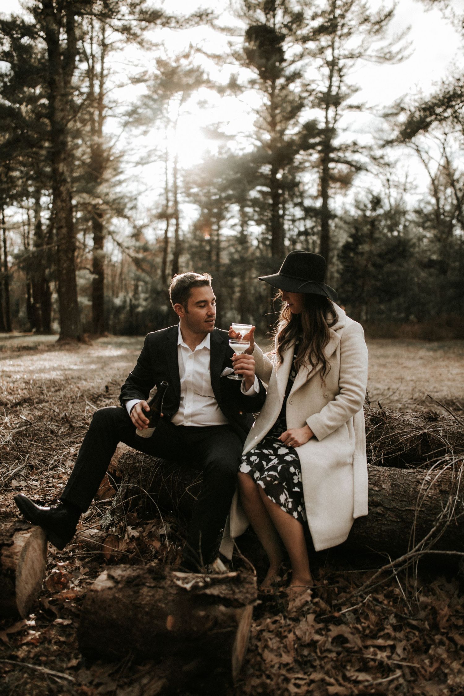Romantic forest photoshoot of couple drinking wine on fallen tree trunk.