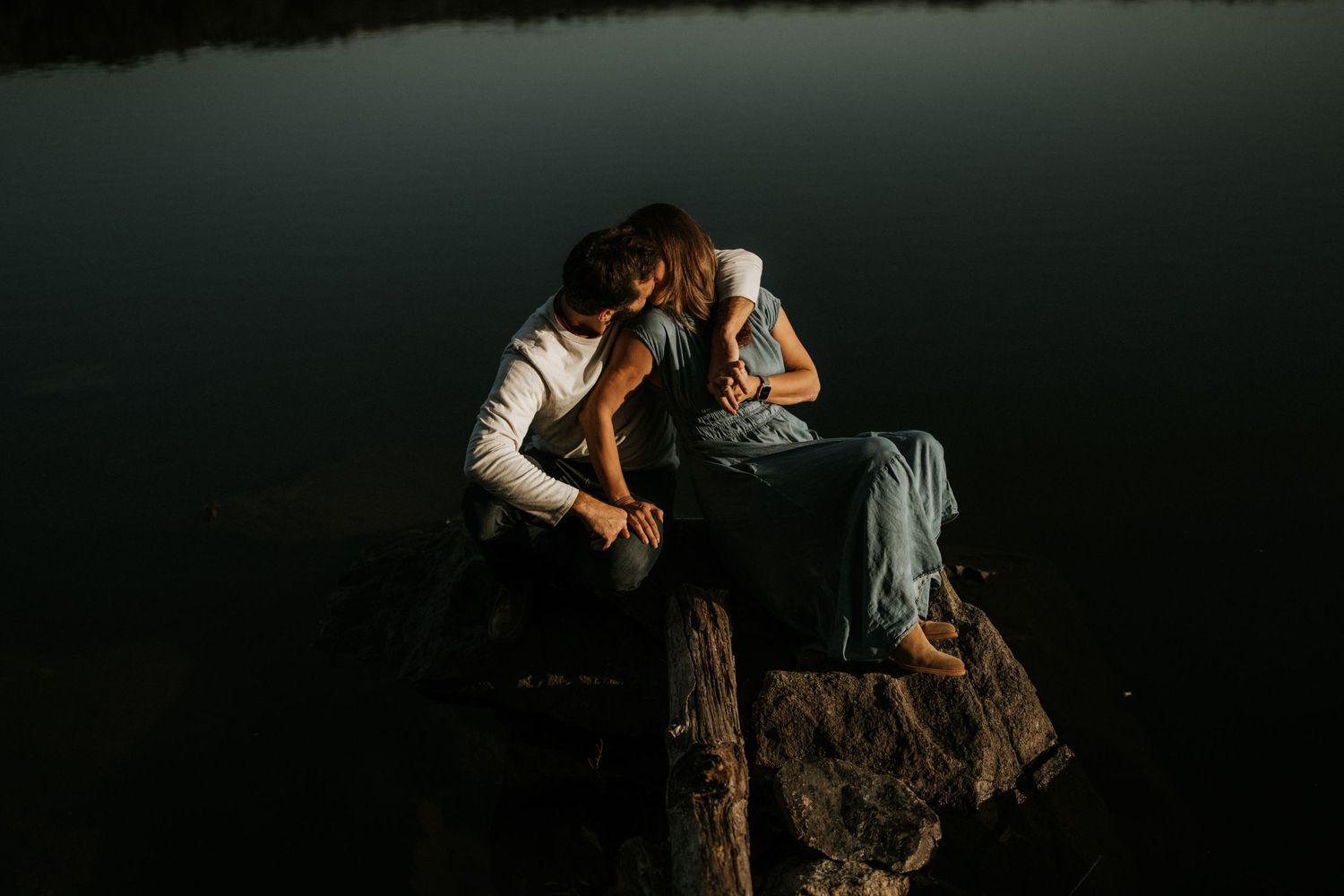 Two people sit close together on rocks during a moody outdoor photoshoot.