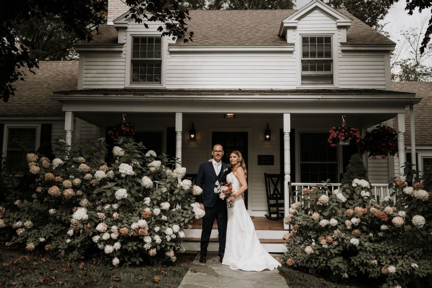 A white farmhouse with a wraparound porch and rose bushes creates a charming wedding venue setting.