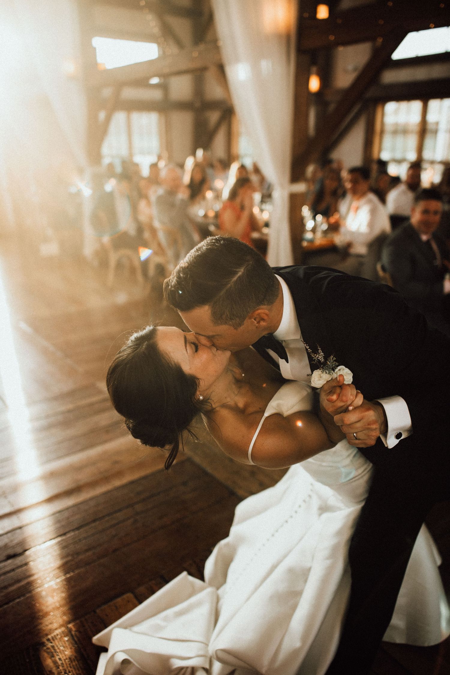 A couple shares their first dance in a sunlit room with wooden beams and guests in the background.