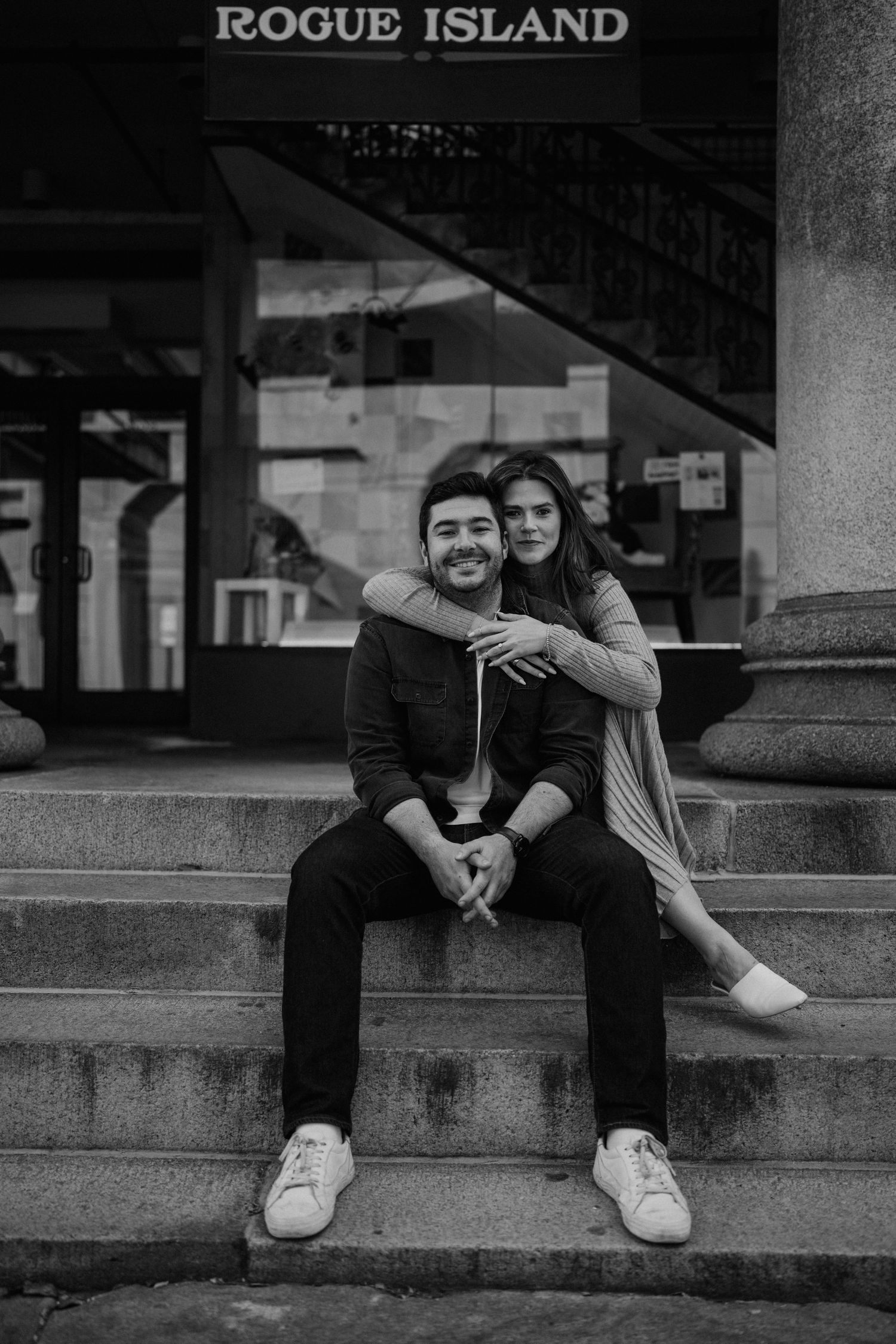 A couple relaxes on concrete steps in front of a Rogue Island sign.