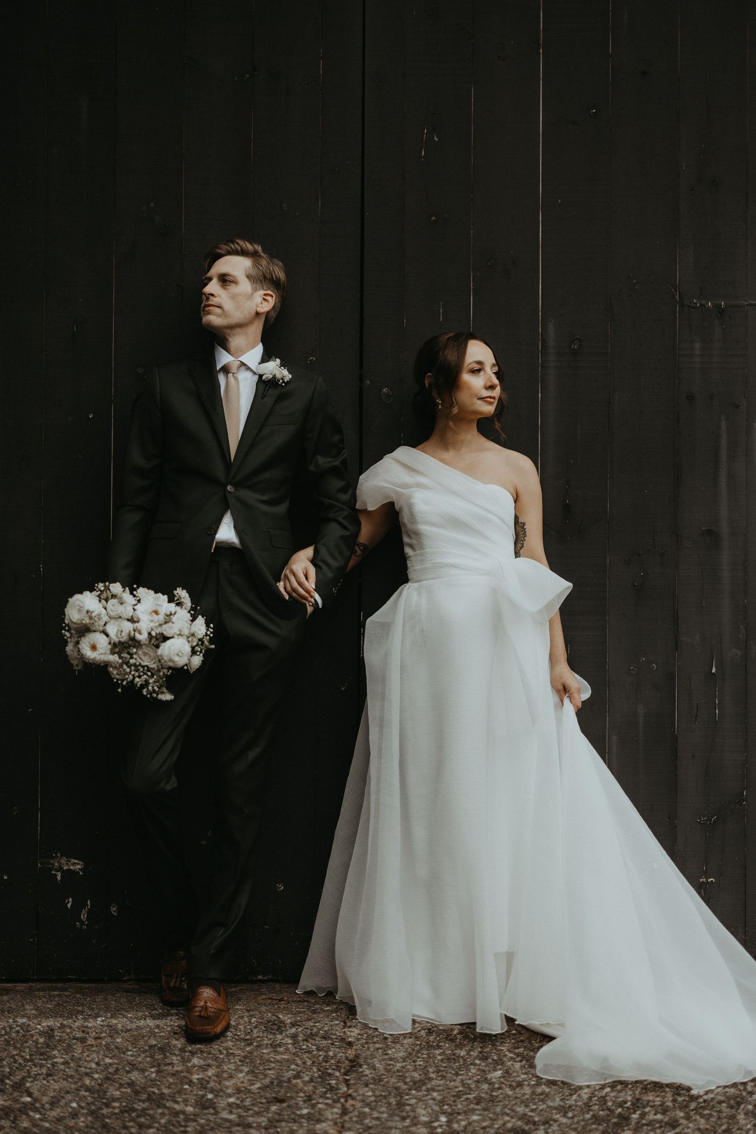 A couple in wedding attire poses against a dark wooden wall with a white floral bouquet.