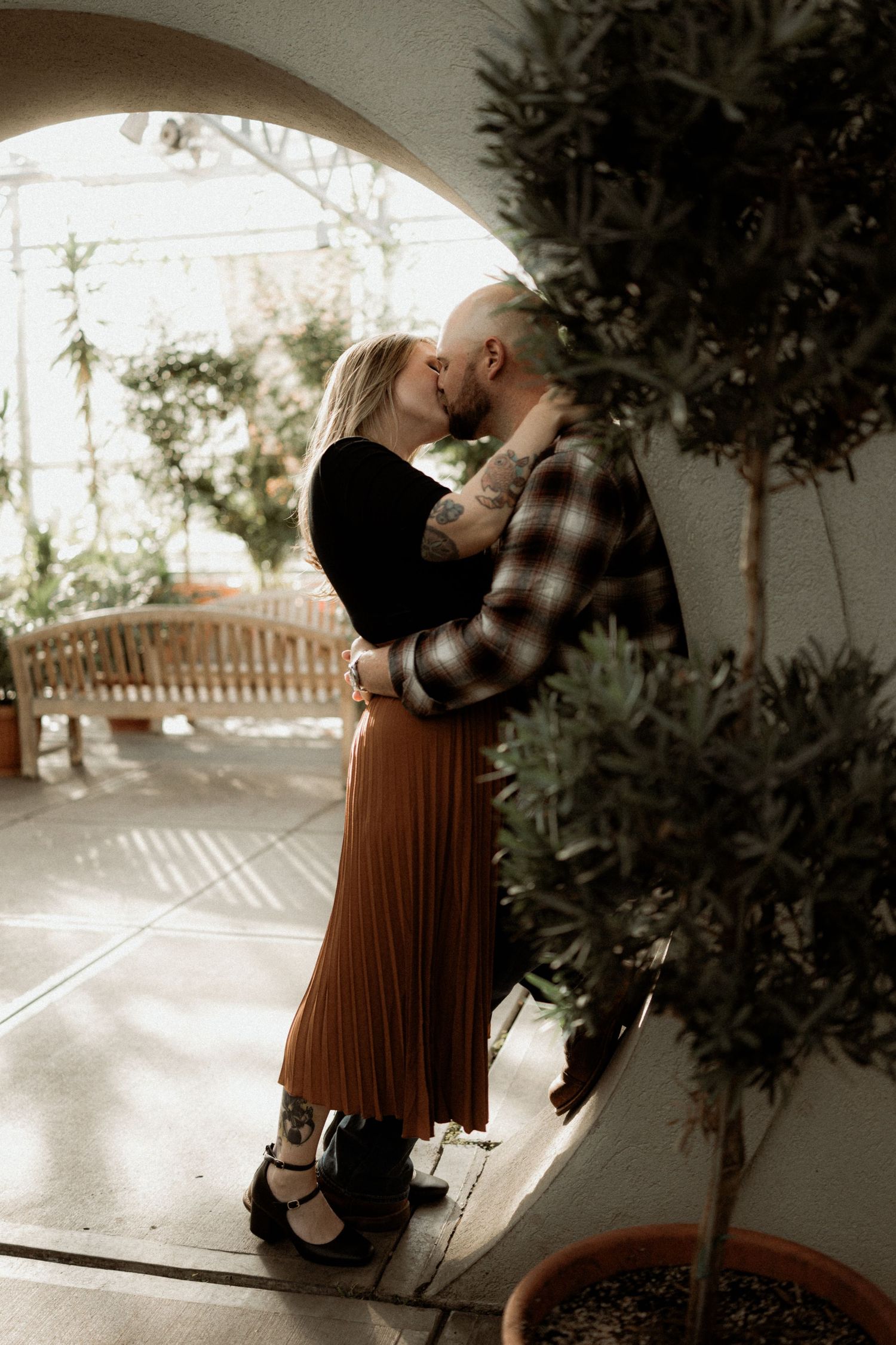 Romantic couple embraces in a sunlit indoor space with potted trees creating an intimate atmosphere.