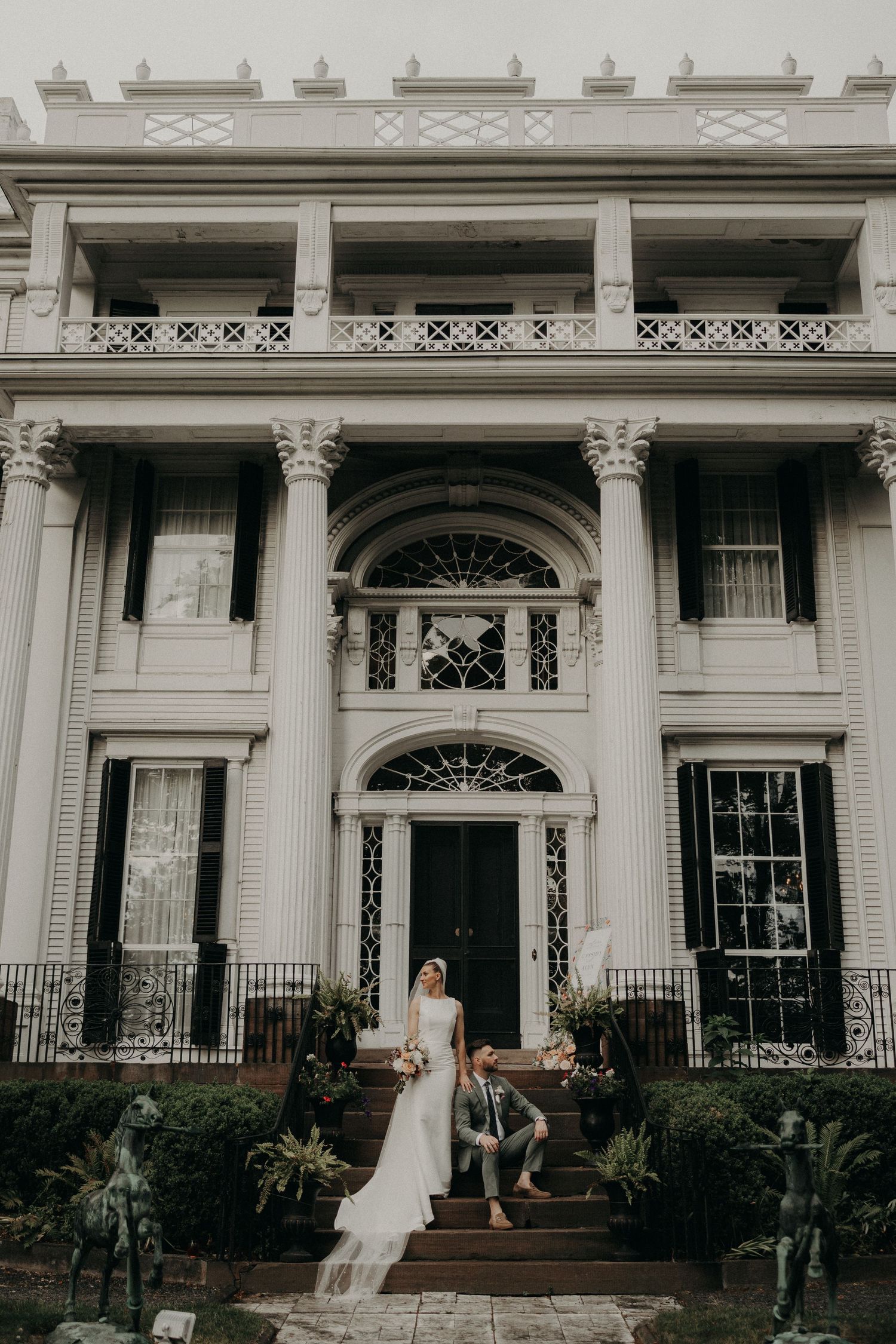 Bride and groom pose on steps of elegant white neoclassical mansion with grand columns and ornate ironwork balcony.
