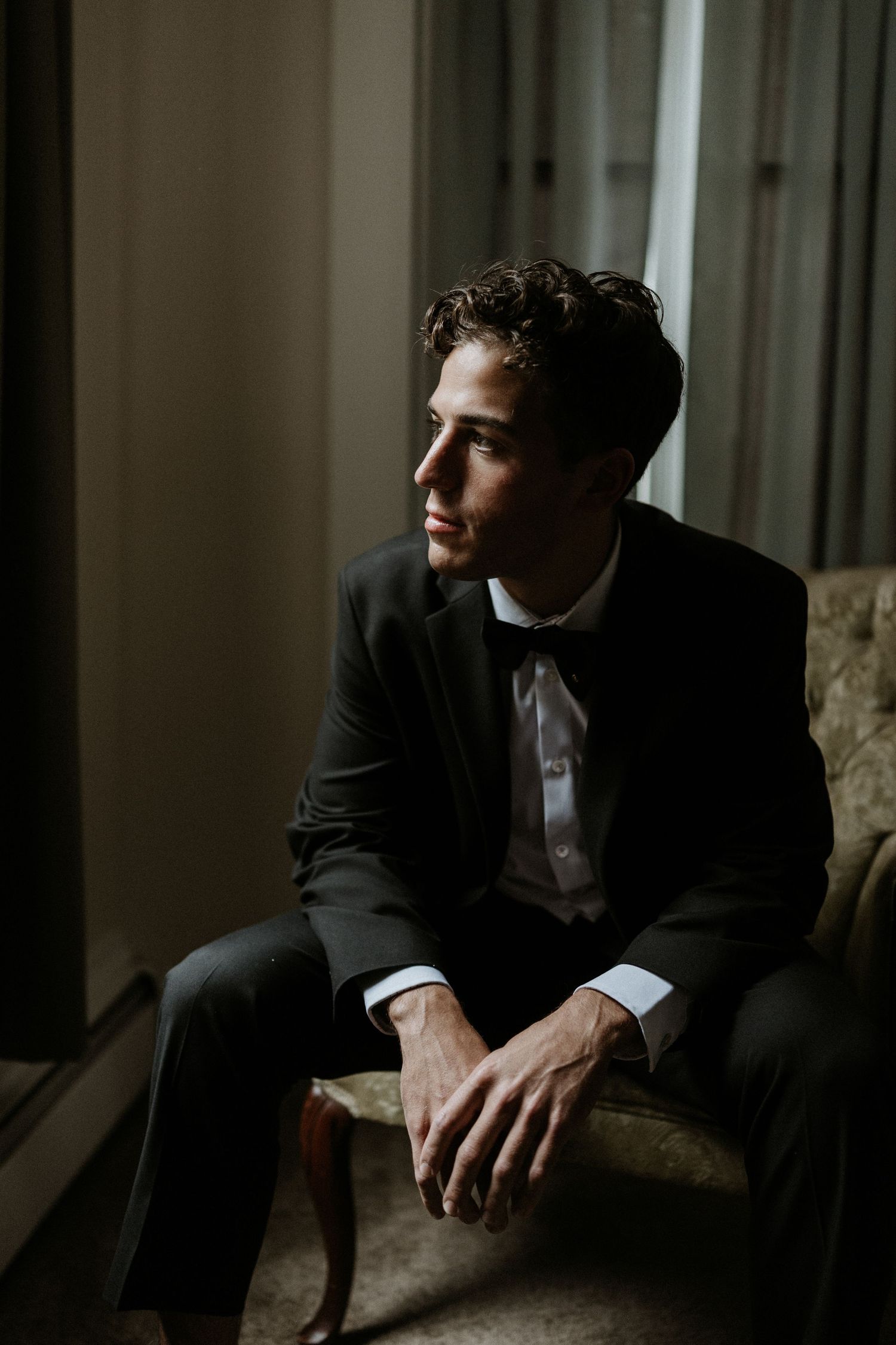A person in a black tuxedo and bowtie sits thoughtfully in a dimly lit room.