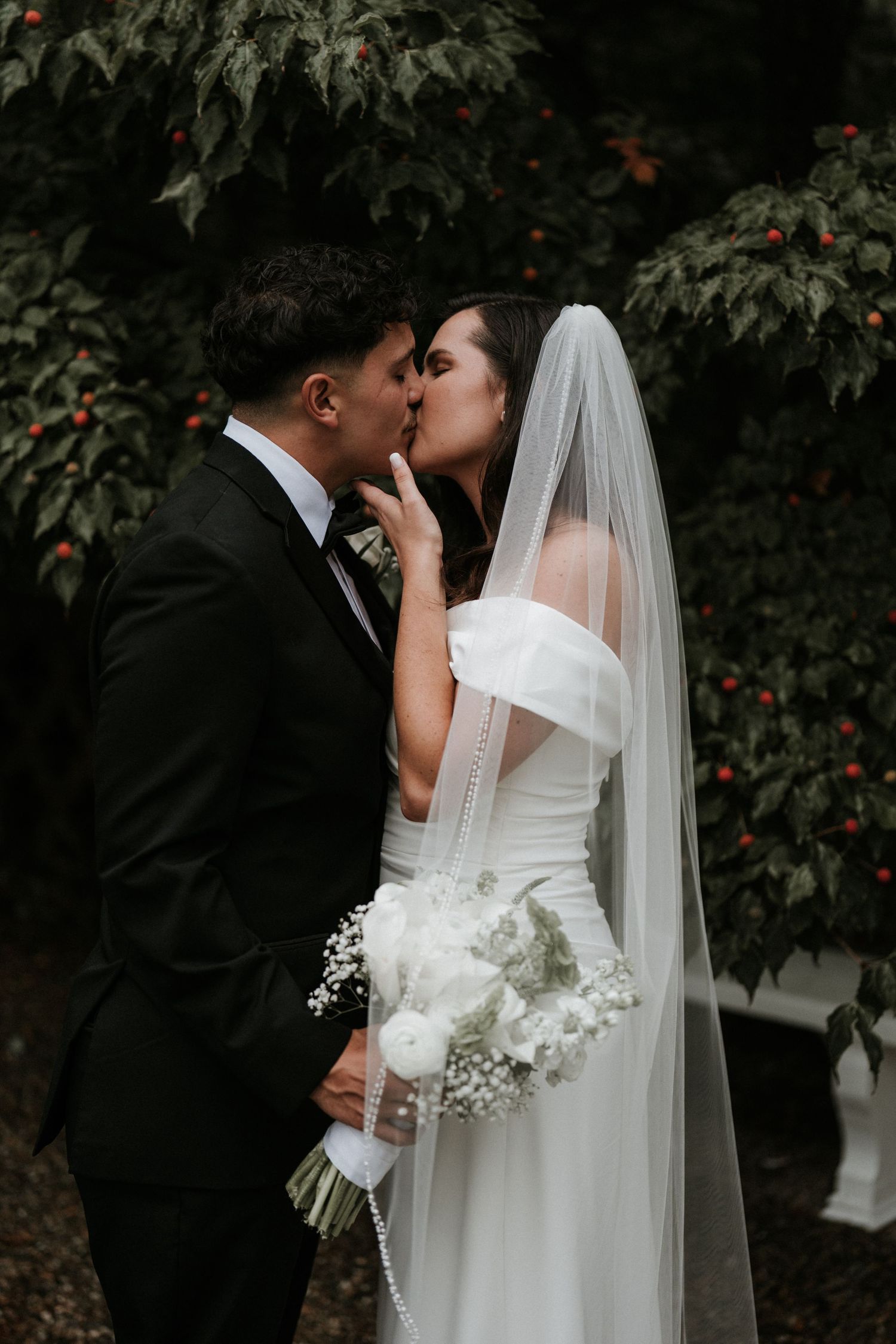 Romantic wedding couple embraces outdoors surrounded by lush greenery and white flowers.