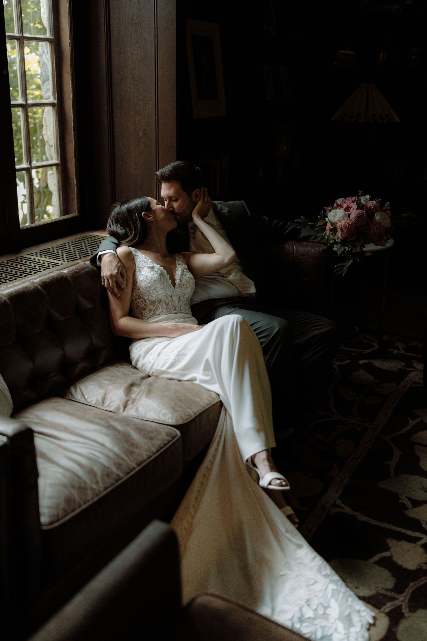 Intimate moment between couple on vintage couch near window in dark moody setting.