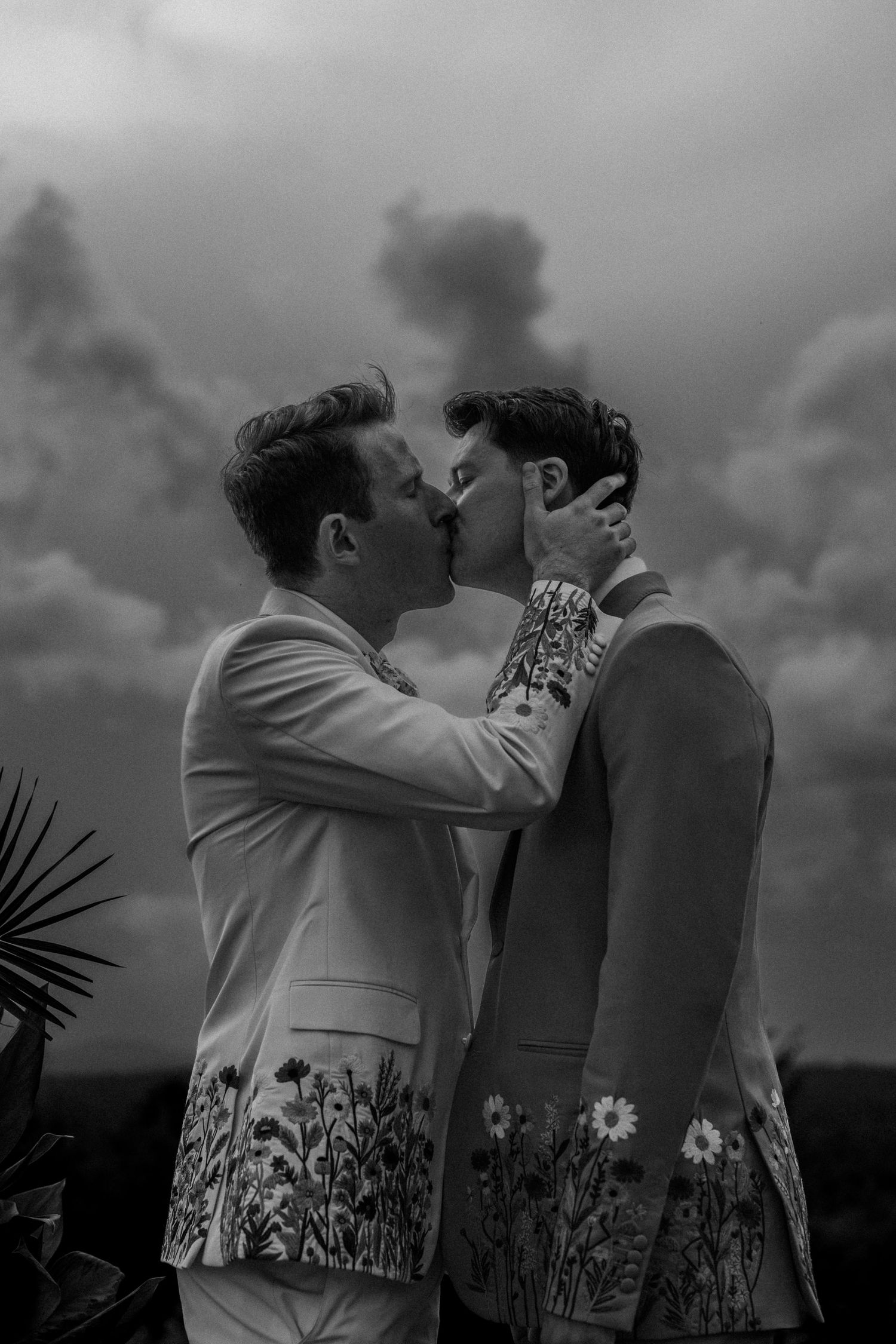 Two grooms in floral-embroidered suits share a romantic kiss against a dramatic cloudy sky in a black and white wedding photo.