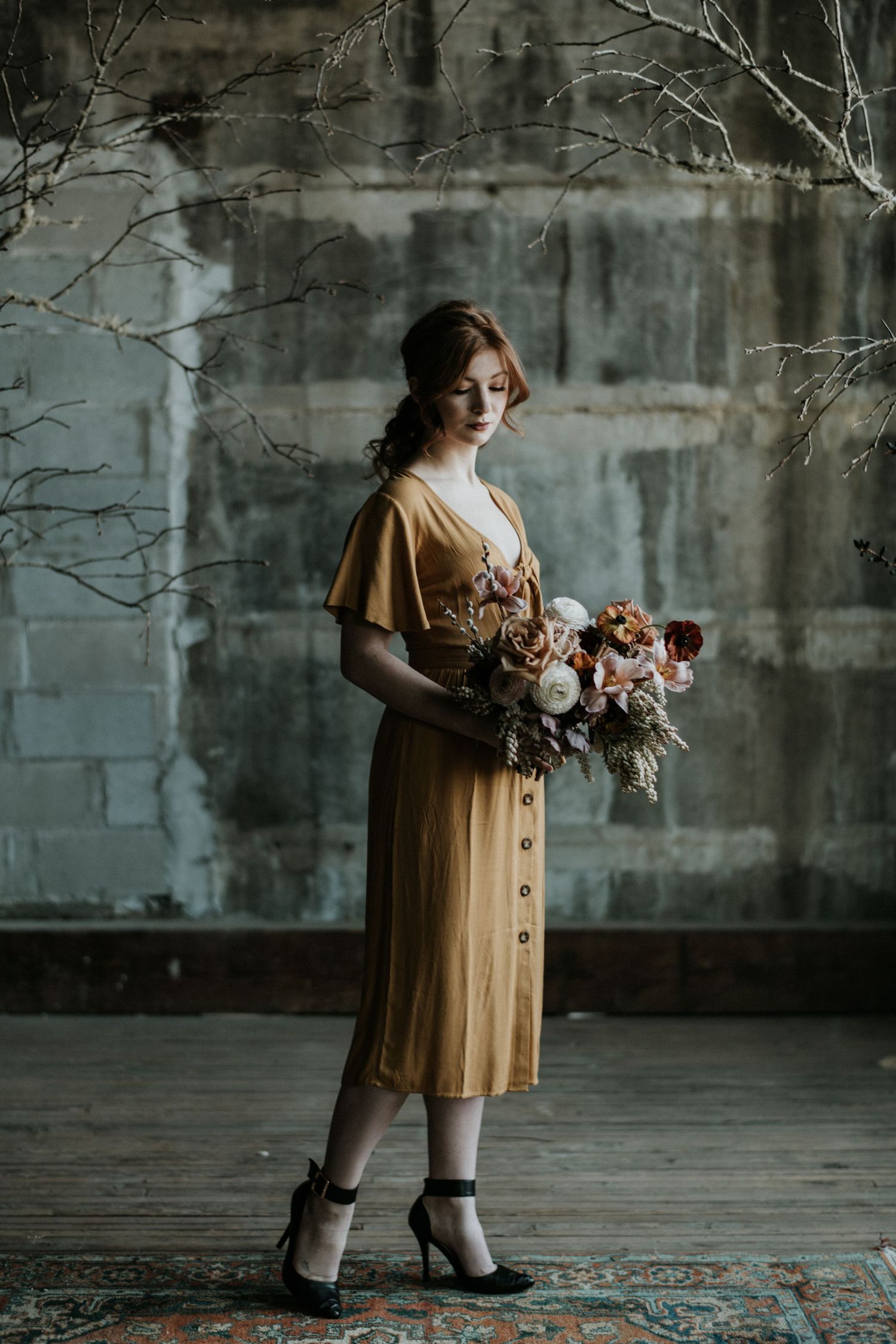 Person in a mustard dress holding flowers against an industrial backdrop.