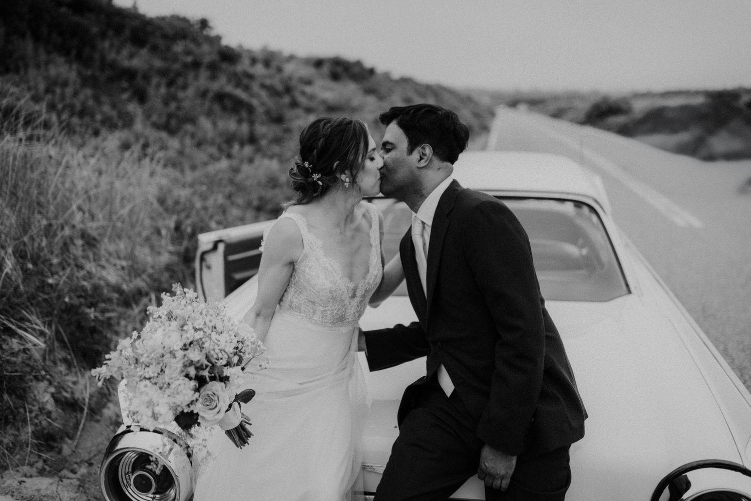 Black and white wedding photo of couple kissing against classic car.