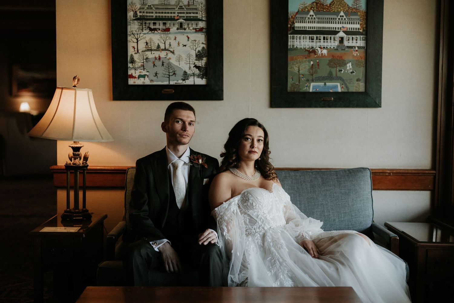 A couple in wedding attire poses together on an elegant blue sofa with framed artwork behind them.