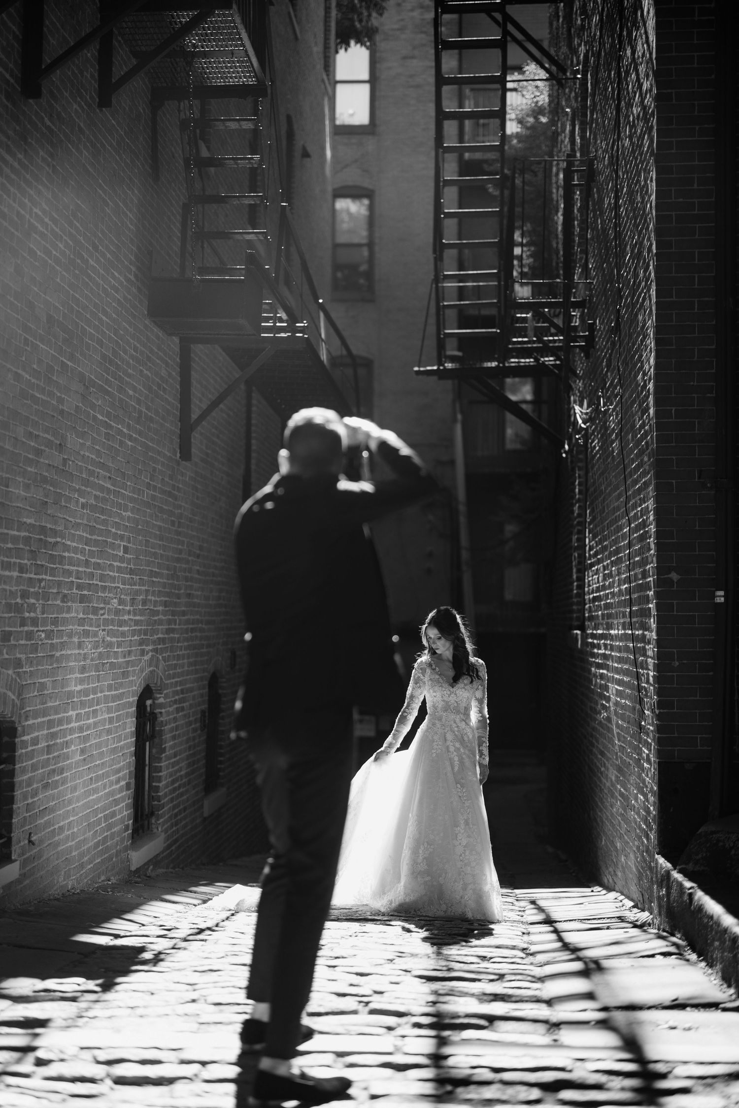 Black and white wedding photo in narrow alley between brick buildings with dramatic lighting.