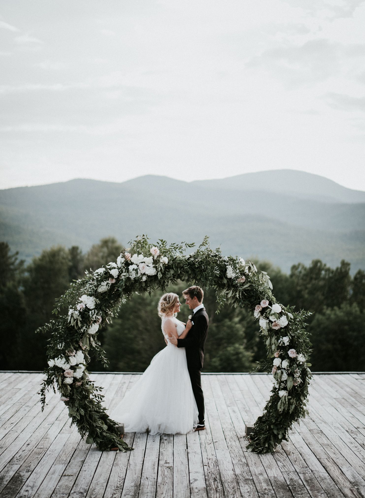 Wedding couple poses between floral arches on deck with mountain landscape backdrop.