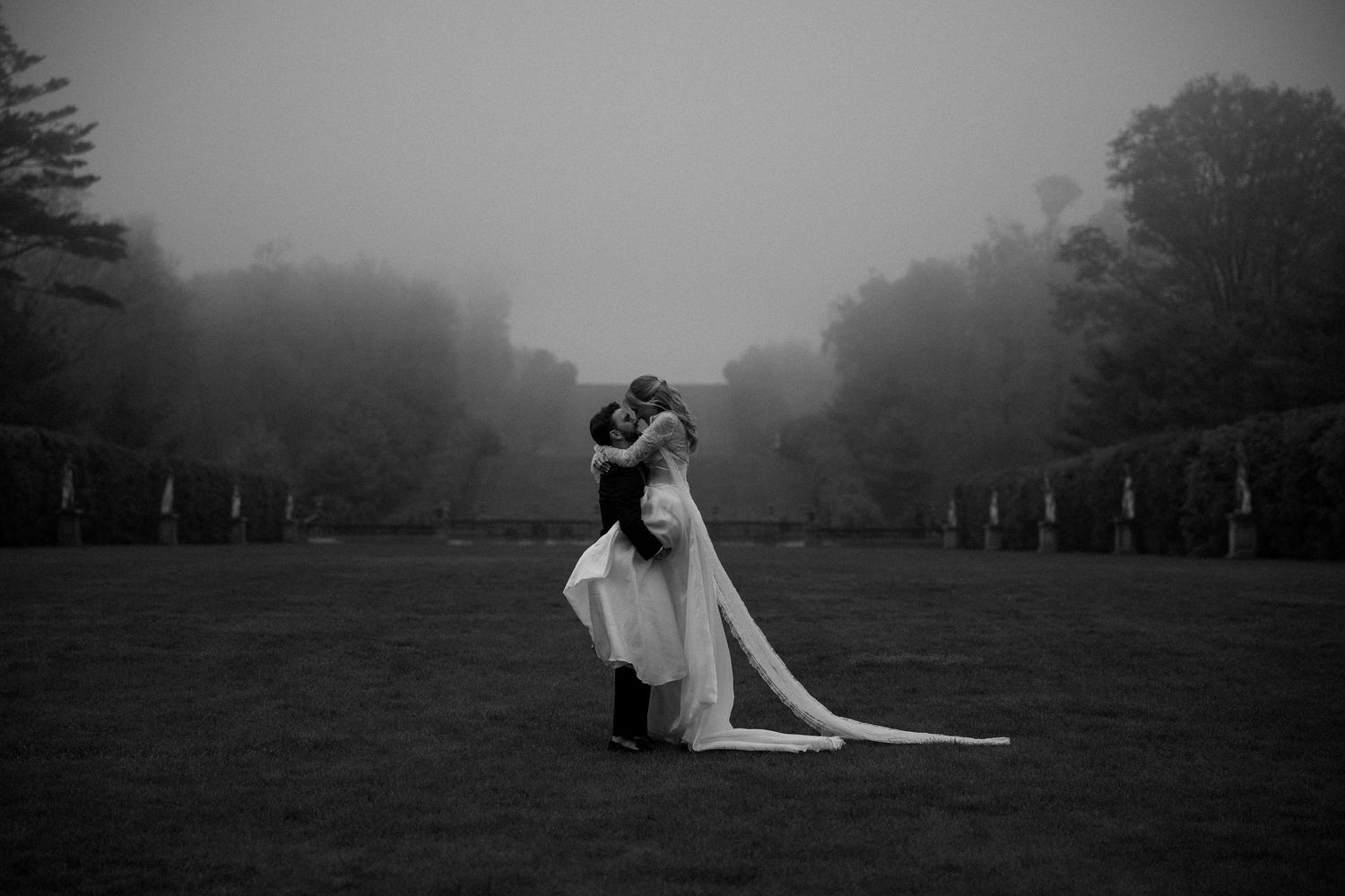 Black and white photo of a couple embracing on a misty lawn, bride in flowing gown, romantic wedding portrait.