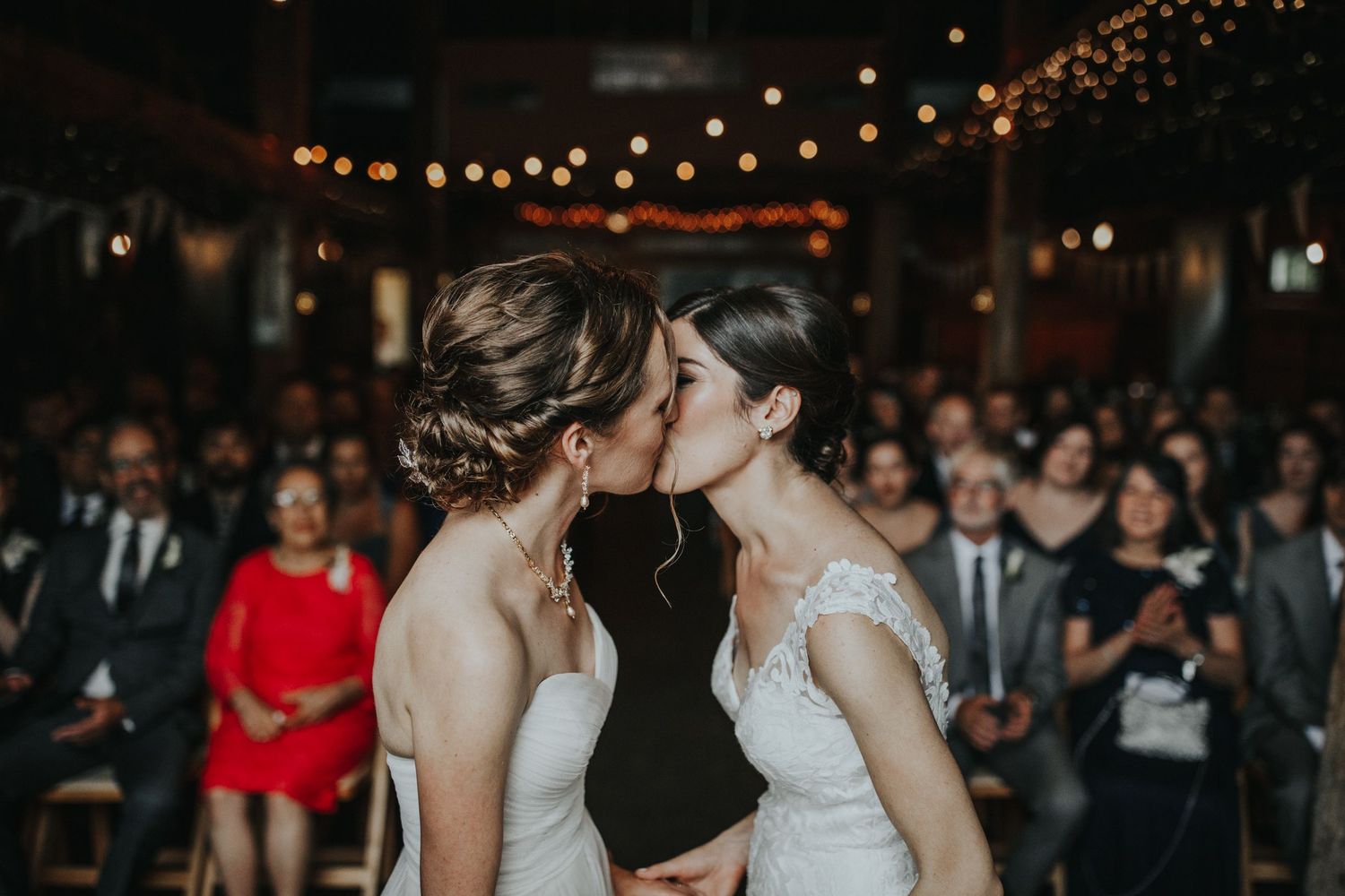 Two brides share an intimate moment on their wedding day under string lights while guests look on.