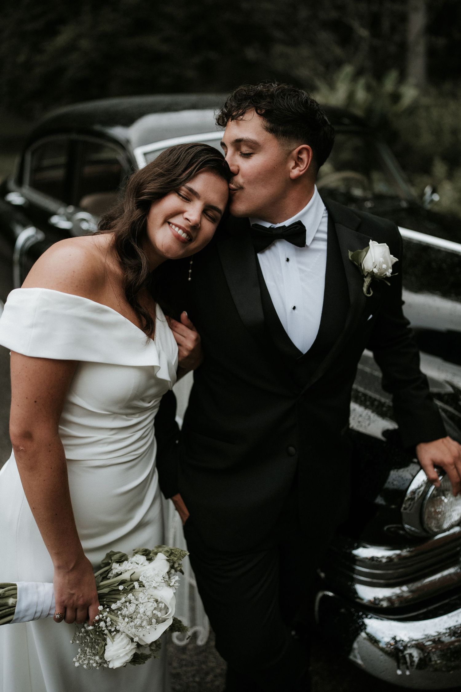 Intimate wedding portraits captured against vintage cars in black and white tones.