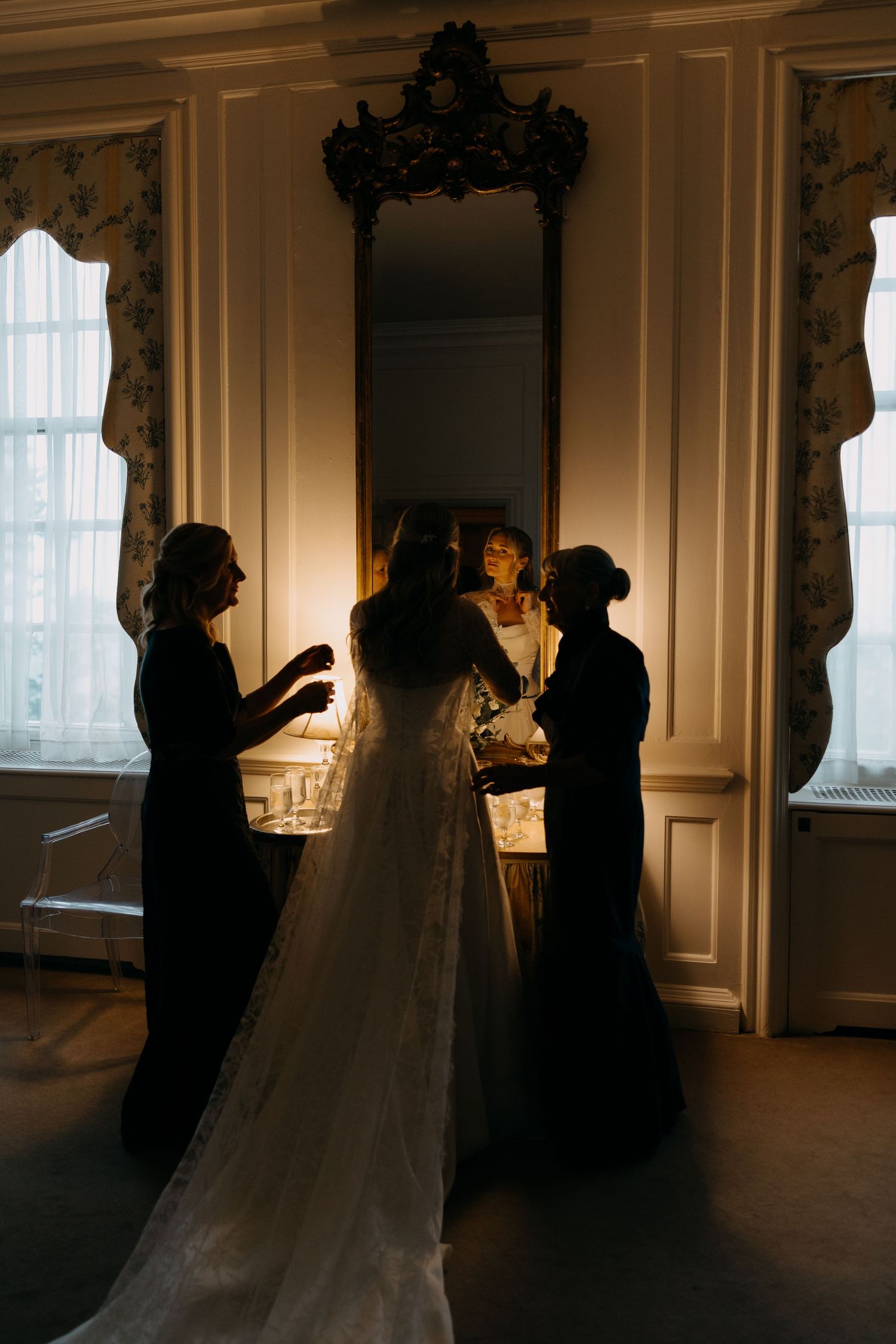 Bride in white gown with long train stands with two women in dark dresses before ornate mirror in elegant room.