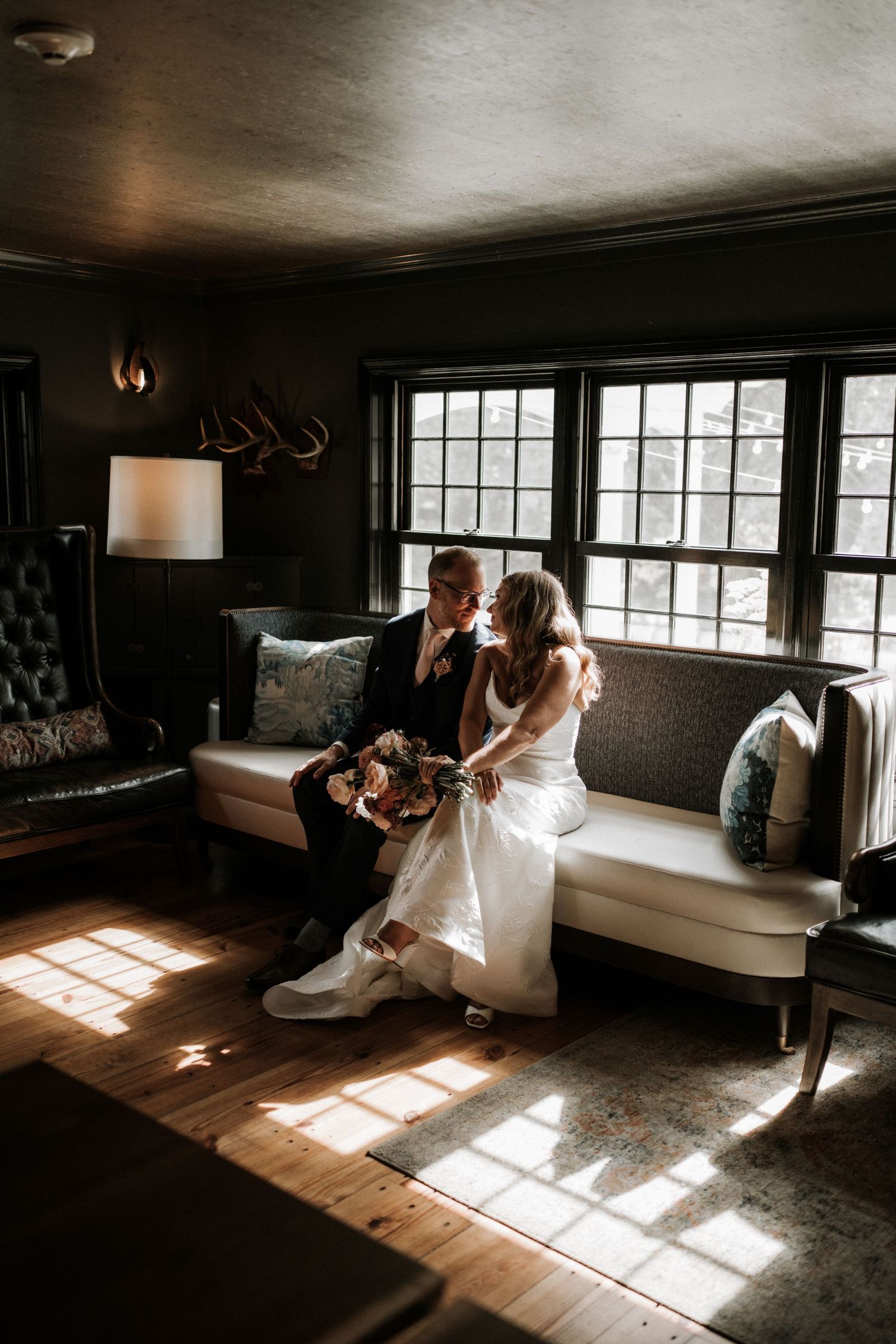 Bride and groom share a romantic moment on a couch in a moody, sunlit room with hardwood floors on their wedding day.