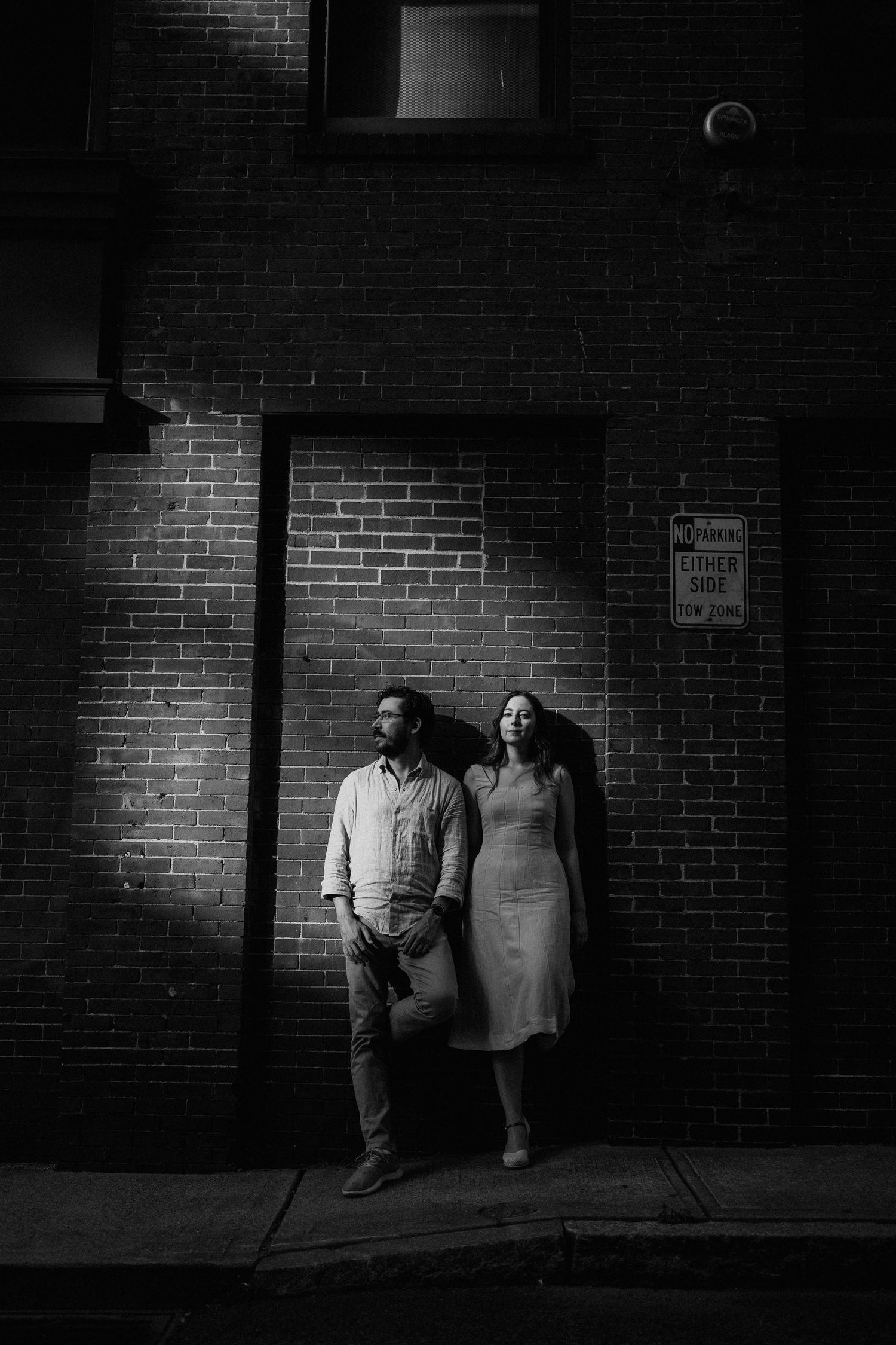 A couple stands against a brick wall in dramatic black and white lighting.