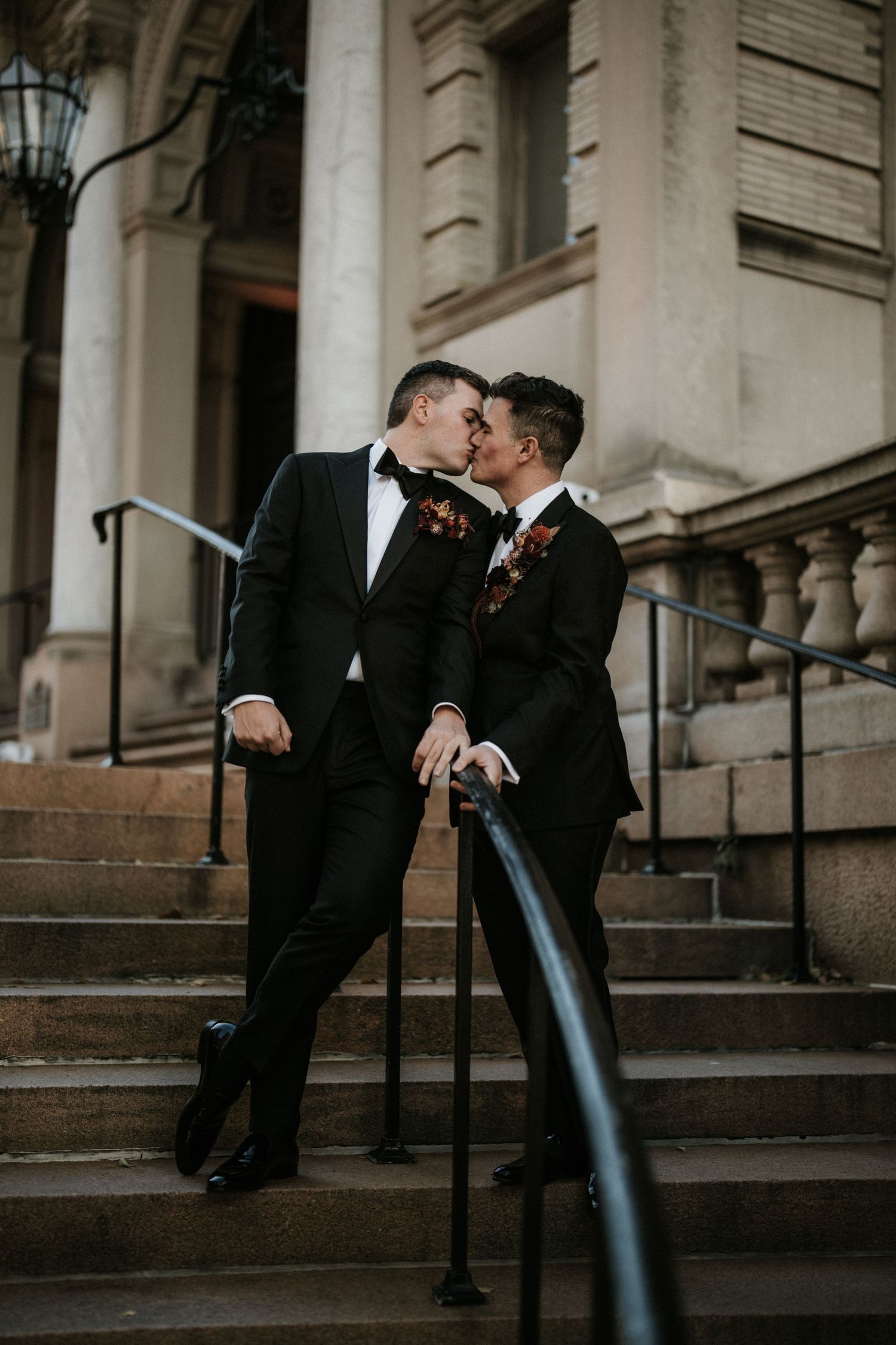 A romantic couple in formal attire shares a tender moment on stone steps outside an elegant building.