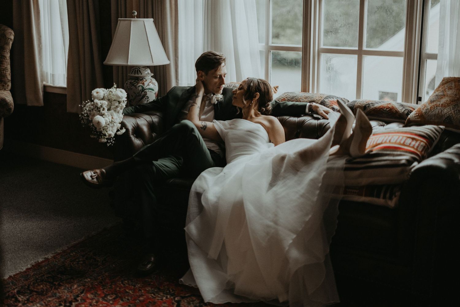 A couple relaxes on a cozy couch in wedding attire with soft window light.