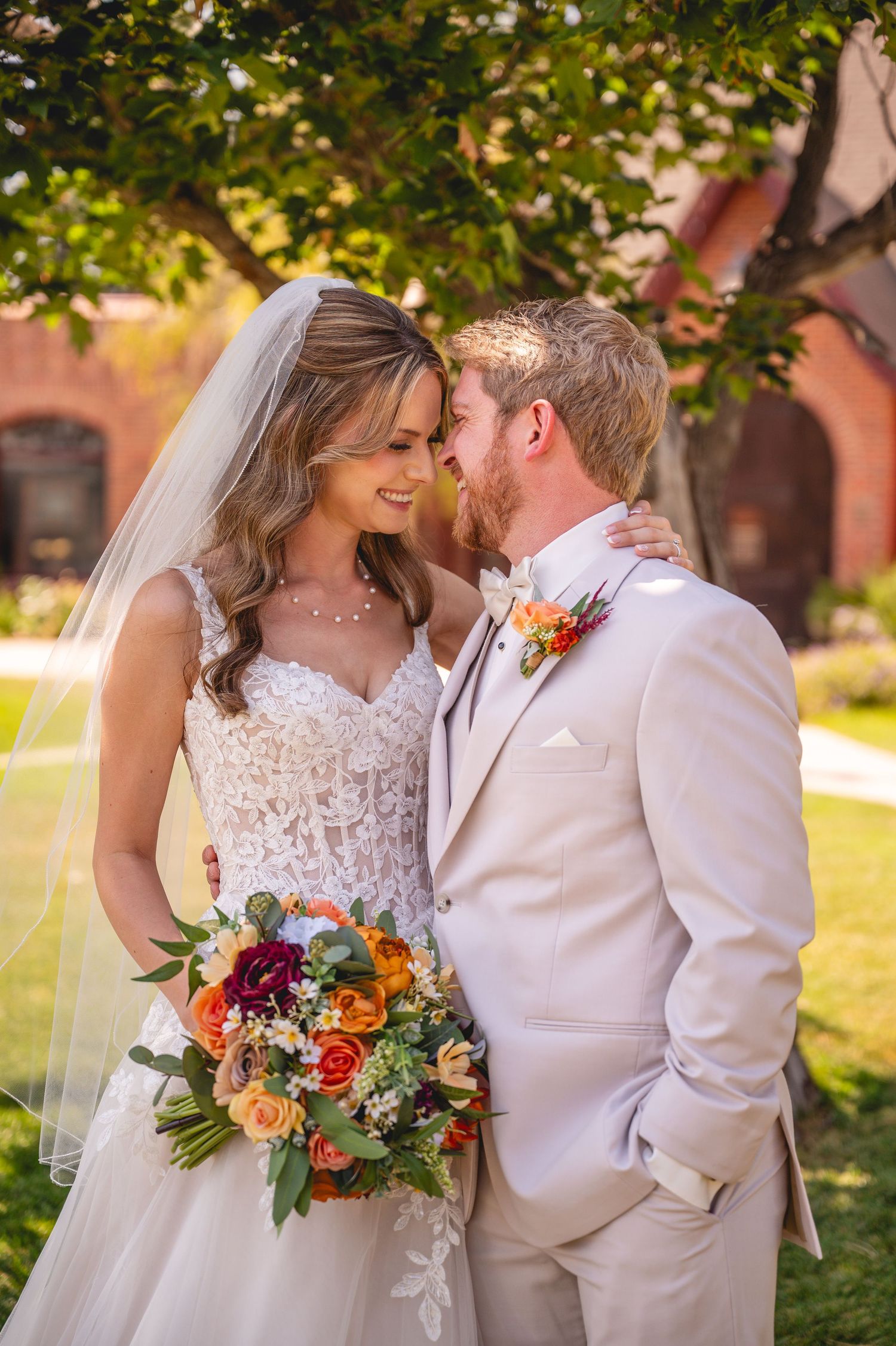 Bride and groom share a romantic moment outdoors, she holds a colorful bouquet, wearing a lace gown and veil.