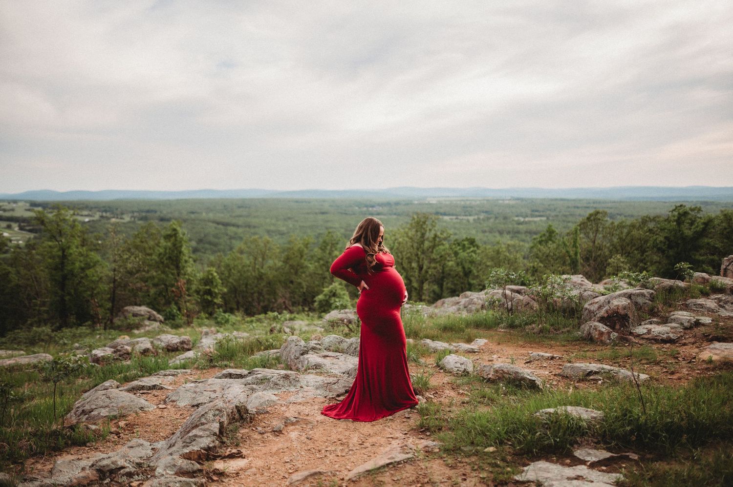 A person in a flowing red dress stands atop a rocky mountain overlook with expansive views of the landscape below.