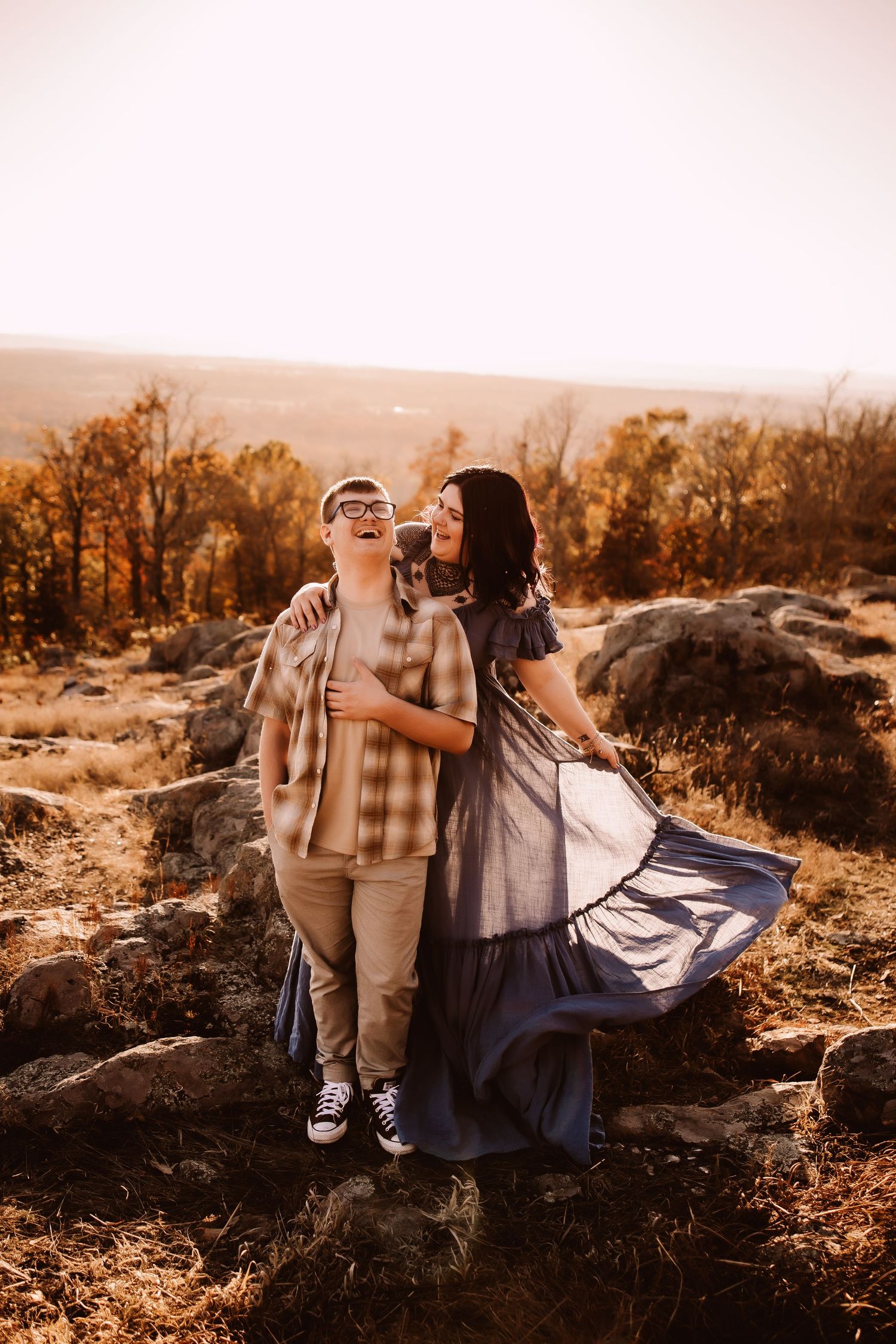 A couple shares a joyful moment at sunset on rocky terrain with autumn trees in the background.