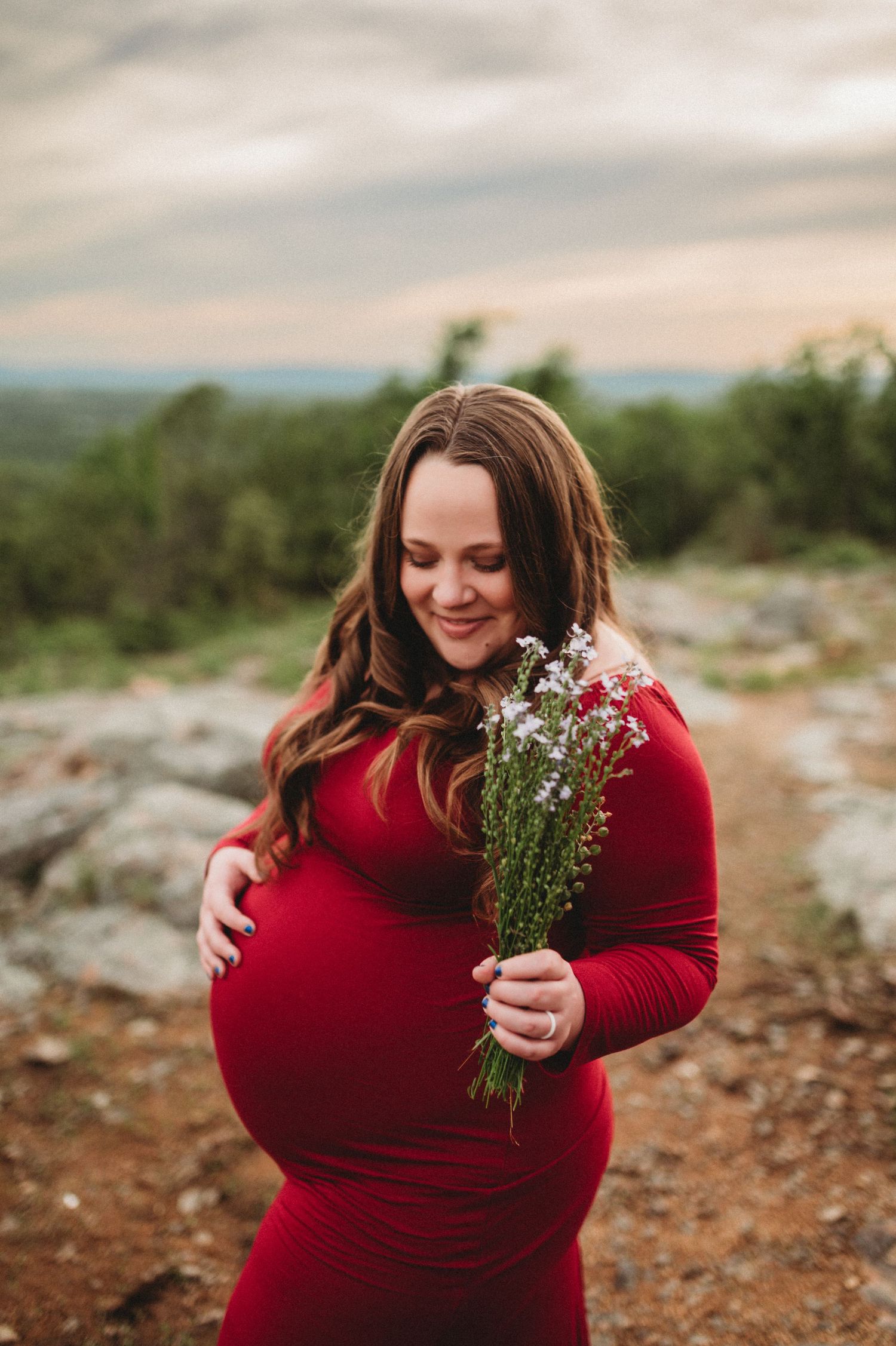 A woman in a red sweater holds wildflowers while posing outdoors during a cloudy day in a natural setting.