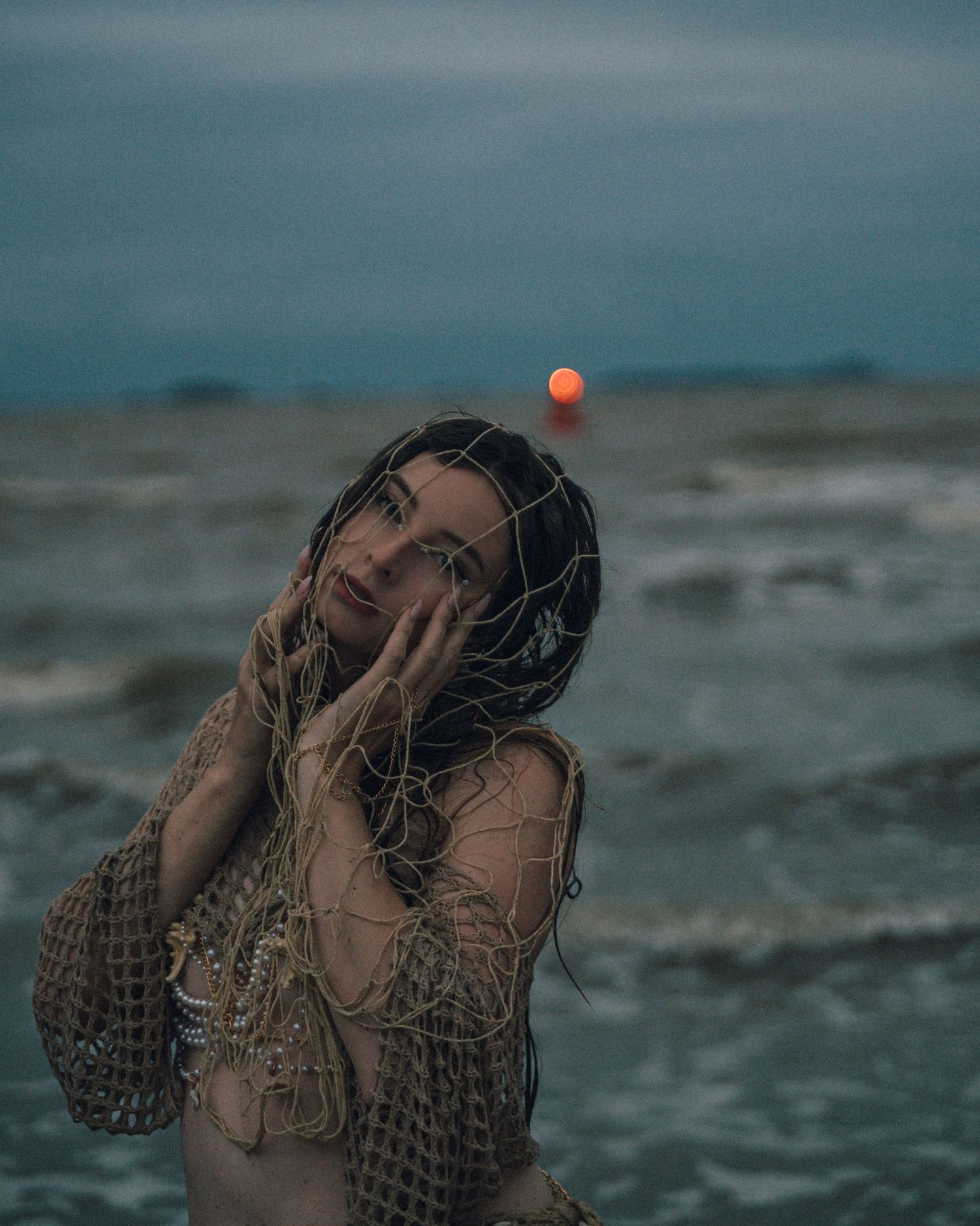 Woman wrapped in a rustic knit net poses in the ocean waves at dusk, with a glowing red sun on the horizon behind her.