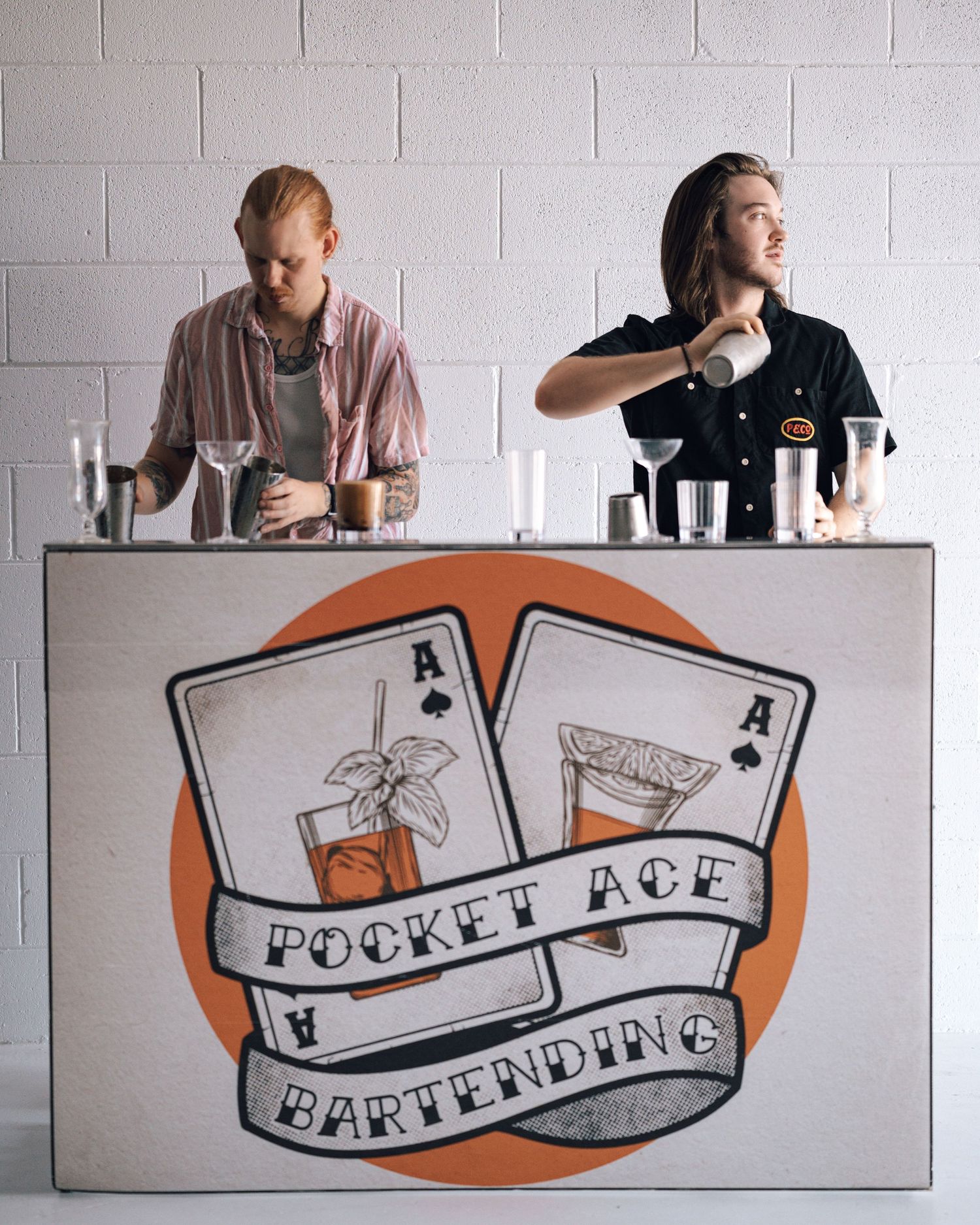 Two bartenders mix drinks behind a Pocket Ace Bartending mobile bar featuring a playing card logo against a white brick wall.