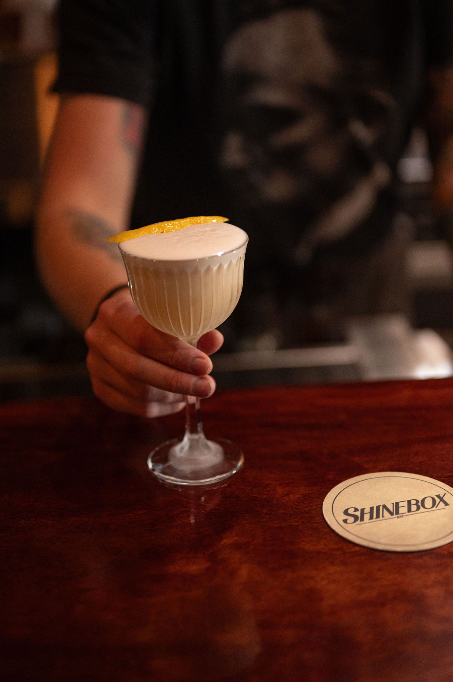 Bartender serves a frothy white cocktail with a lemon twist in a coupe glass on a polished wooden bar beside a Shinebox coaster.