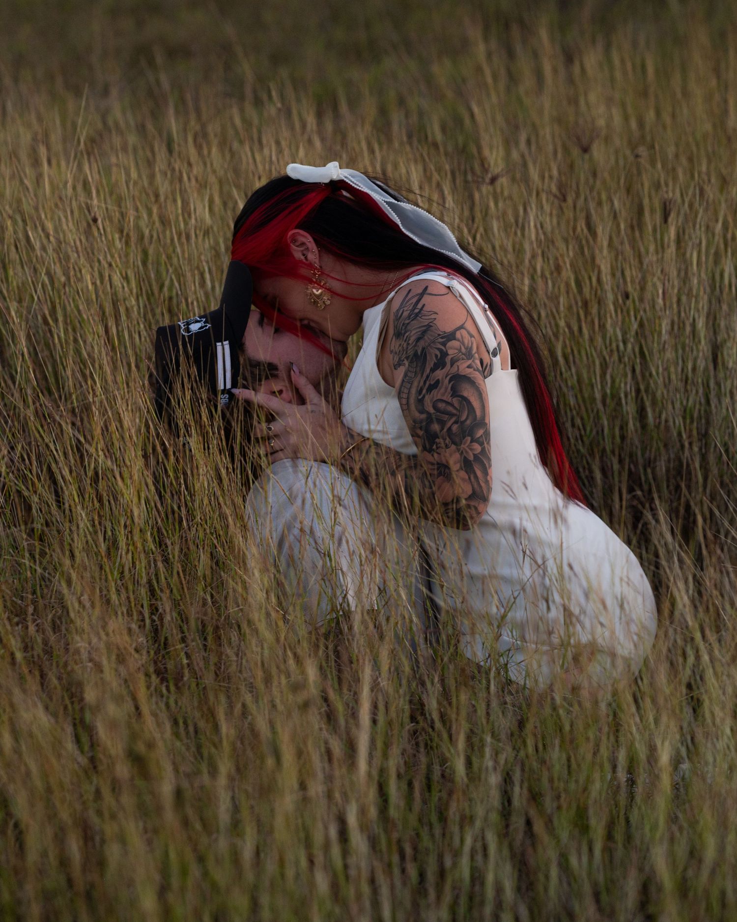 A tattooed woman with red-tipped braids crouches in tall golden grass while holding a camera to her face.